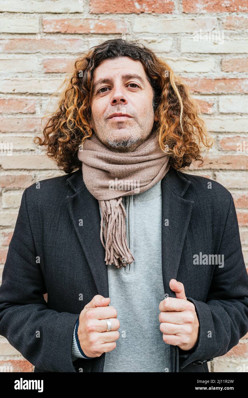 Confident mature man in scarf with curly hair adjusting coat and looking at camera while standing near brick wall Stock Photo