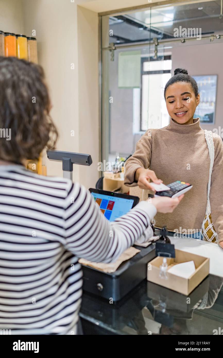 African american woman with eco string bag making payment with credit card on payment terminal in sustainable local shop standing before counter Stock Photo