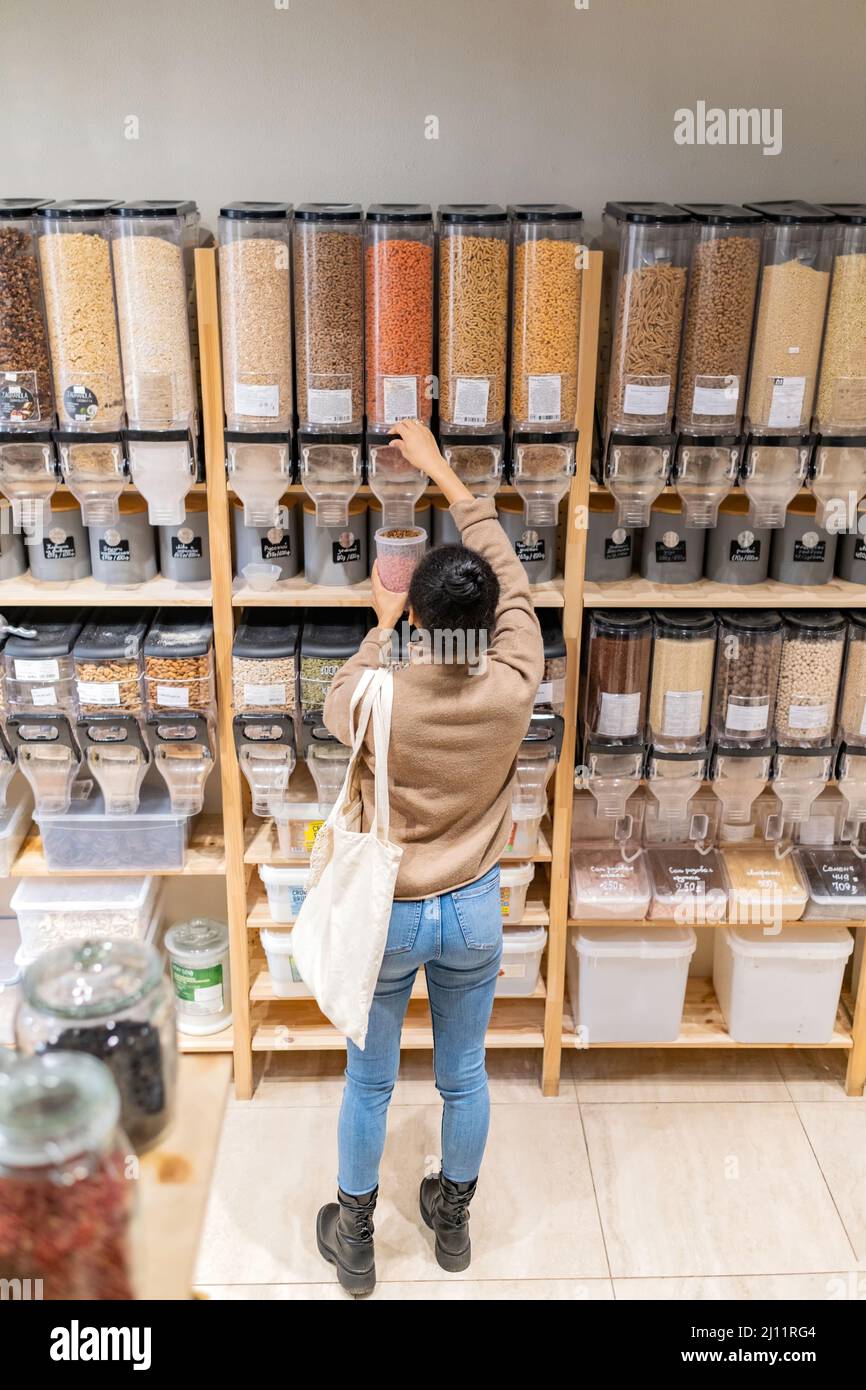 Young african american woman buying gluten free pasta in sustainable zero waste grocery store. Young woman refilling reusable container with pasta in Stock Photo