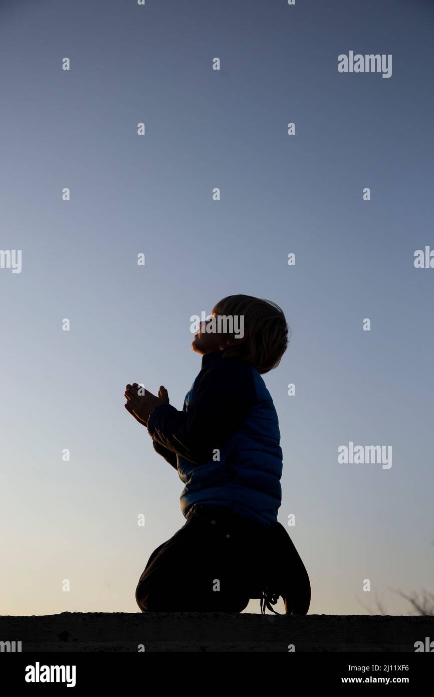 silhouette of a boy kneeling, folded his hands in prayer, an appeal to God. Children against war. A Ukrainian child asks for peace for his homeland Uk Stock Photo