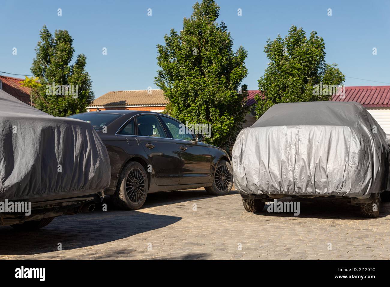 Many cars parked near suburban home driveway covered with protective tarpaulin cloth cover to protect from direct sun light, dust, rain or hail on hot Stock Photo