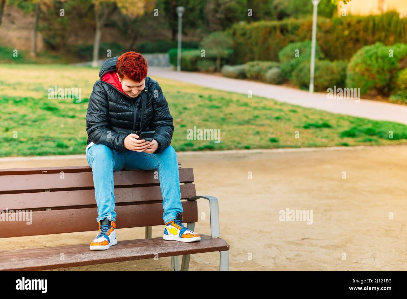 General shot of a modern woman with short red hair, non-binary, looking at her social networks in a public park. Stock Photo