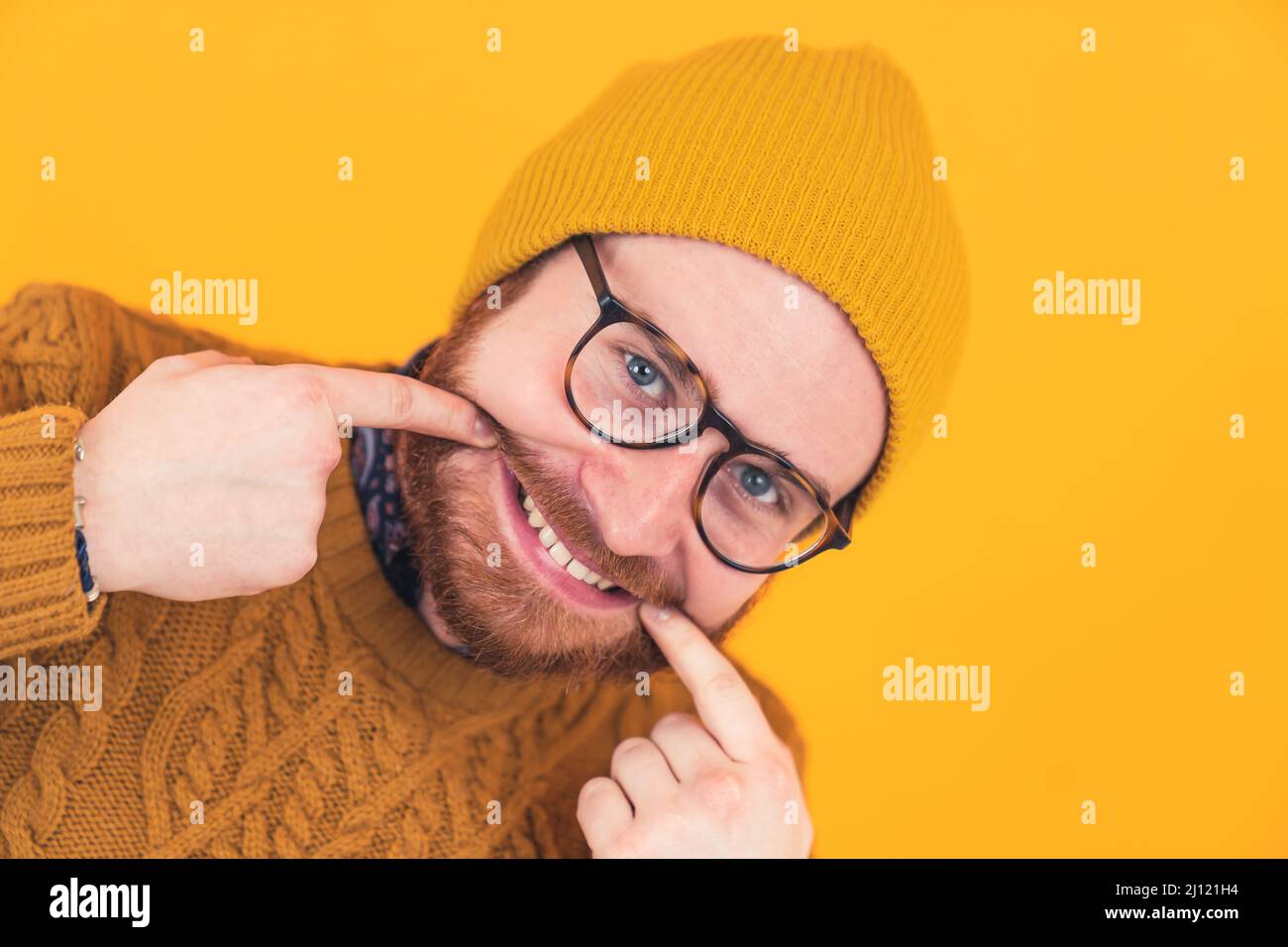 Closeup of cheerful young bearded Caucasian man smiling in glasses and hat, man touching the edges of his mouth, showing teeth orange background isolated studio shot . High quality photo Stock Photo