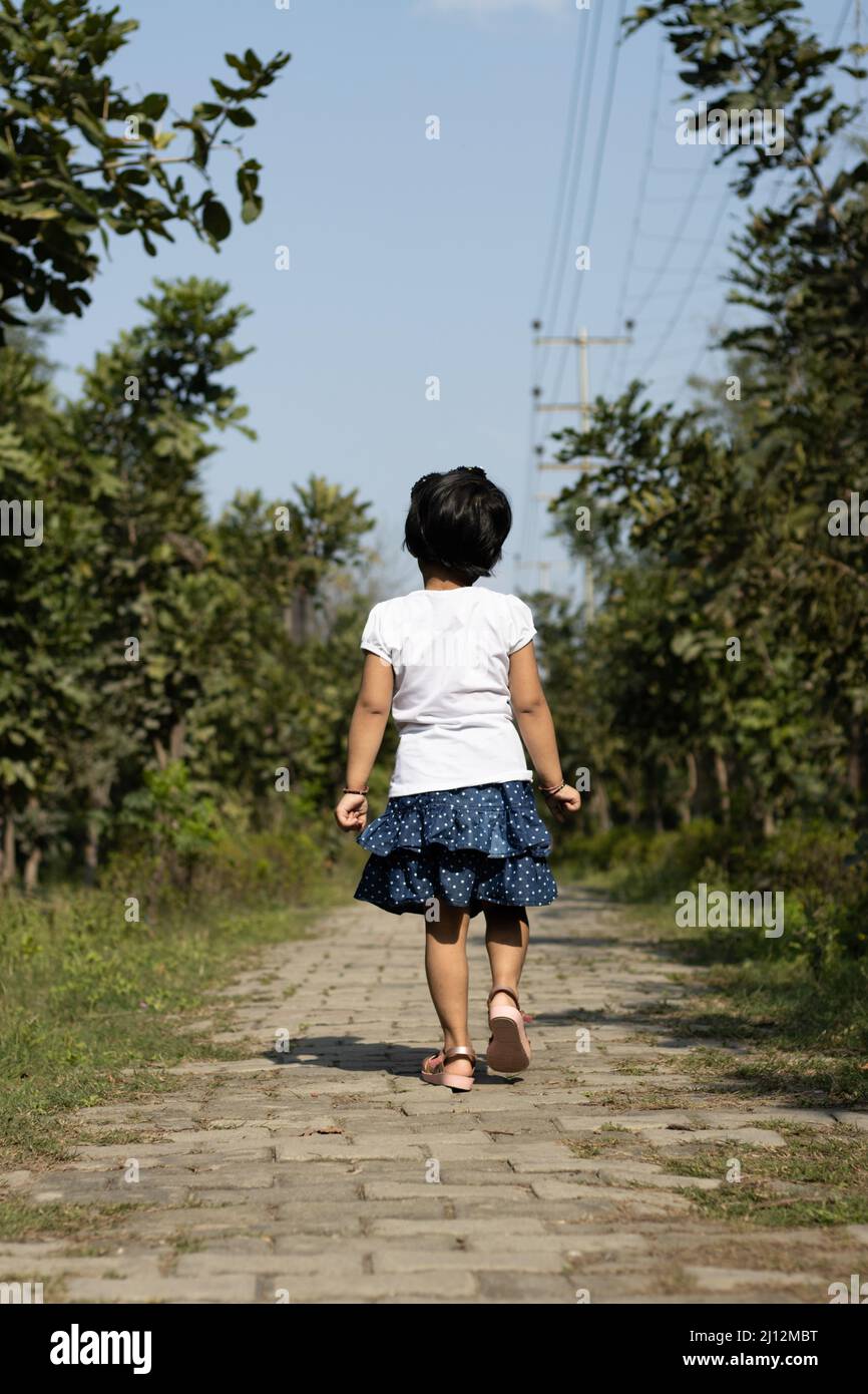 Portrait Of Young Indian Preschooler Girl Kid Walking Away Alone On Parkway In Natural Green Park Stock Photo