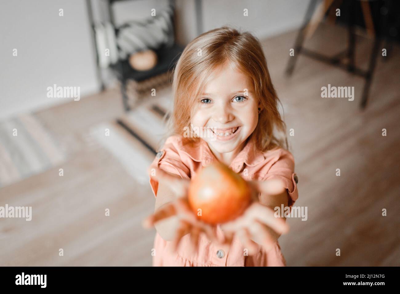 a small cute blonde girl with long hair laughs merrily at the camera and holds an Apple in her hands Stock Photo