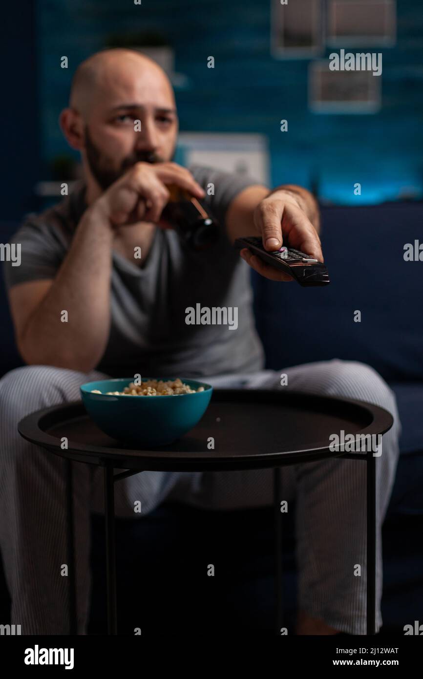 Man drinking beer and searching movie on television, holding TV remote control to switch channel programs. Person watching film or broadcast show with electronic media keypad. Close up. Stock Photo