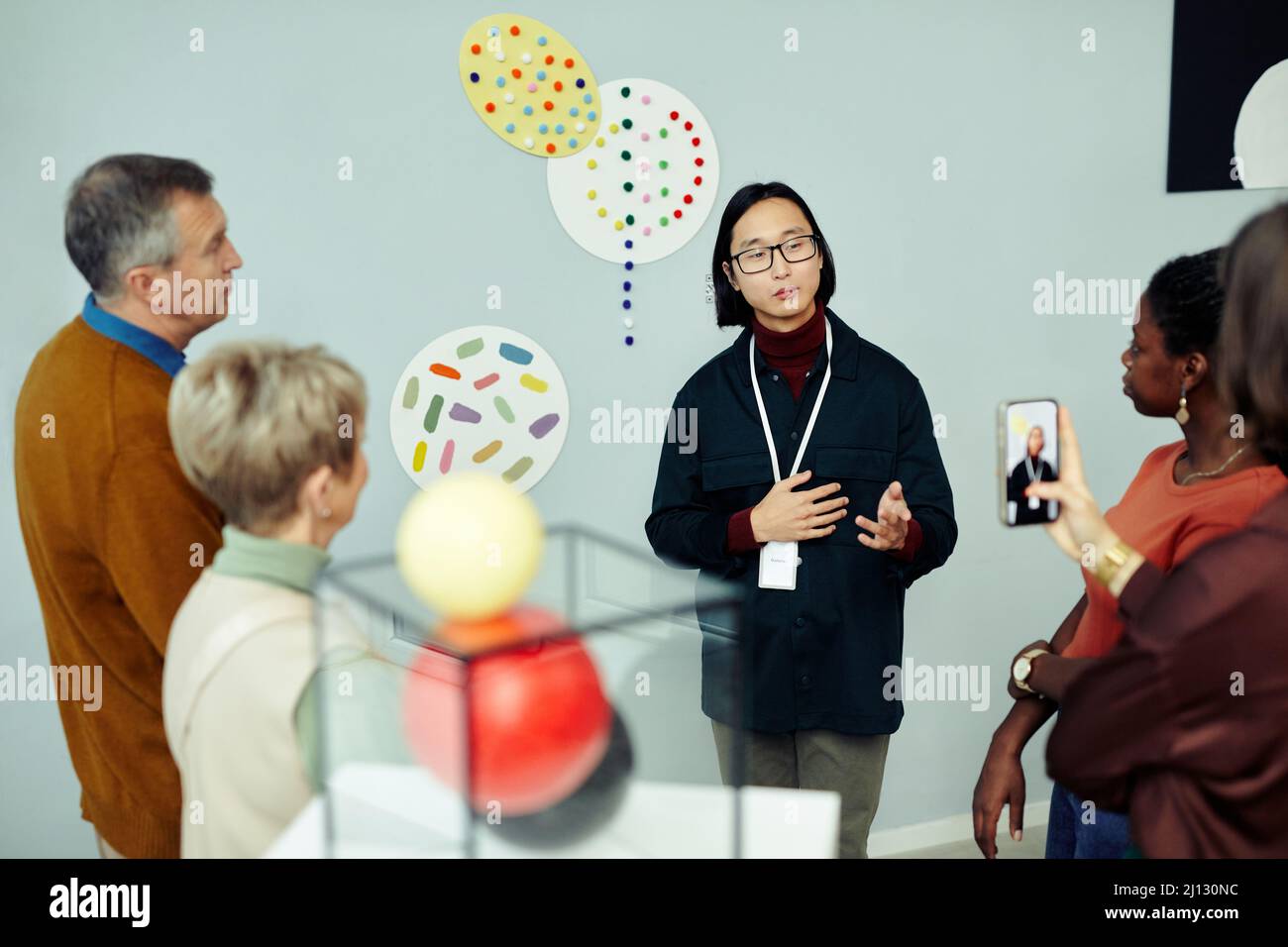 Professional gallery worker standing in front of group of tourists speaking about contemporary paintings and objects displayed at exhibition Stock Photo