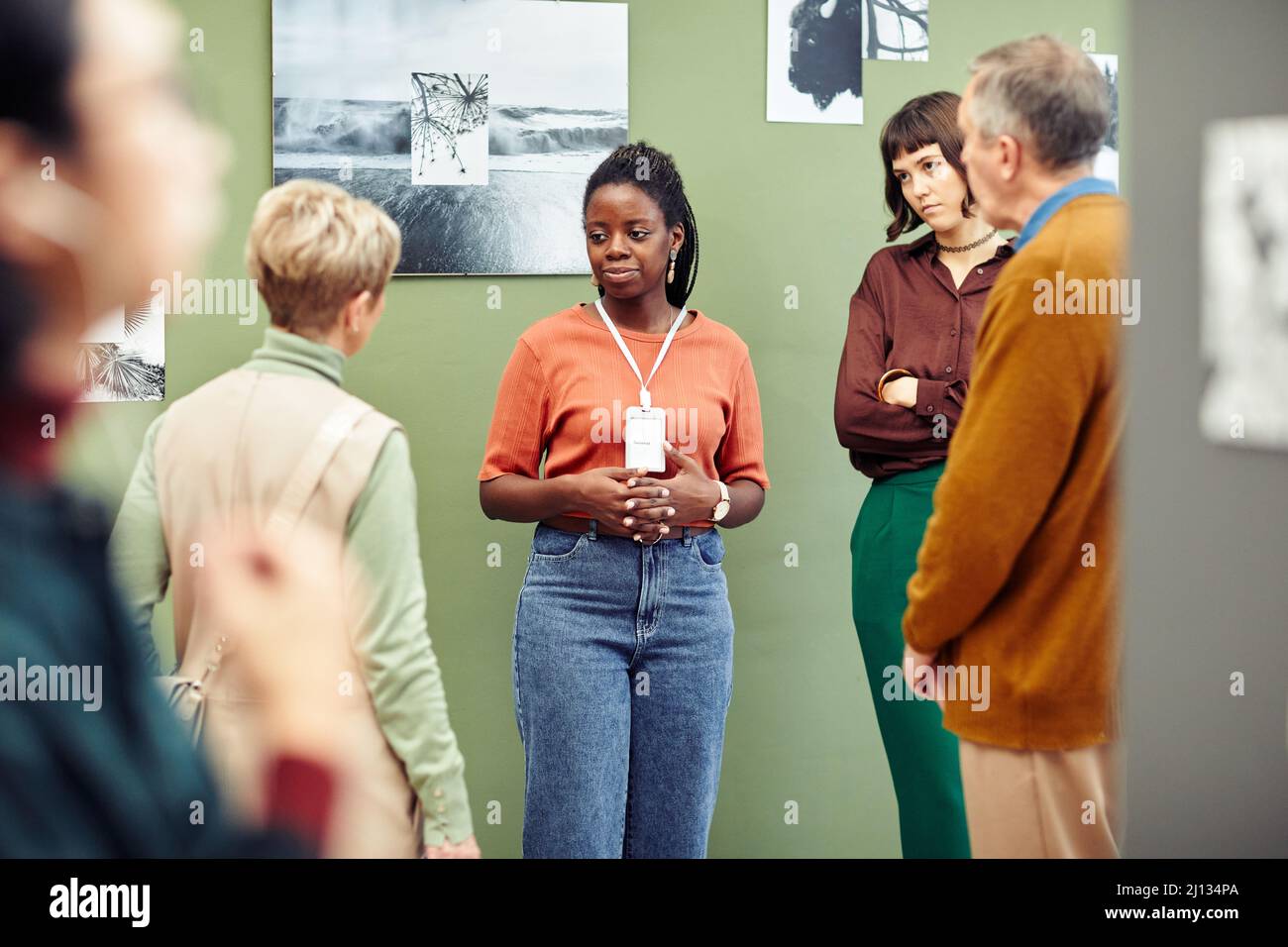Portrait of young African American gallery worker standing in front of visitors discussing black and white photography Stock Photo