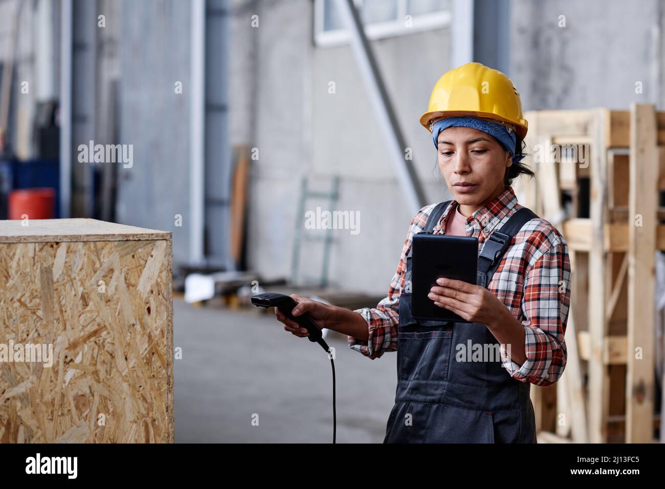 Young serious woman in coveralls and hardhat scanning barcode on huge box and looking at data on screen of tablet Stock Photo