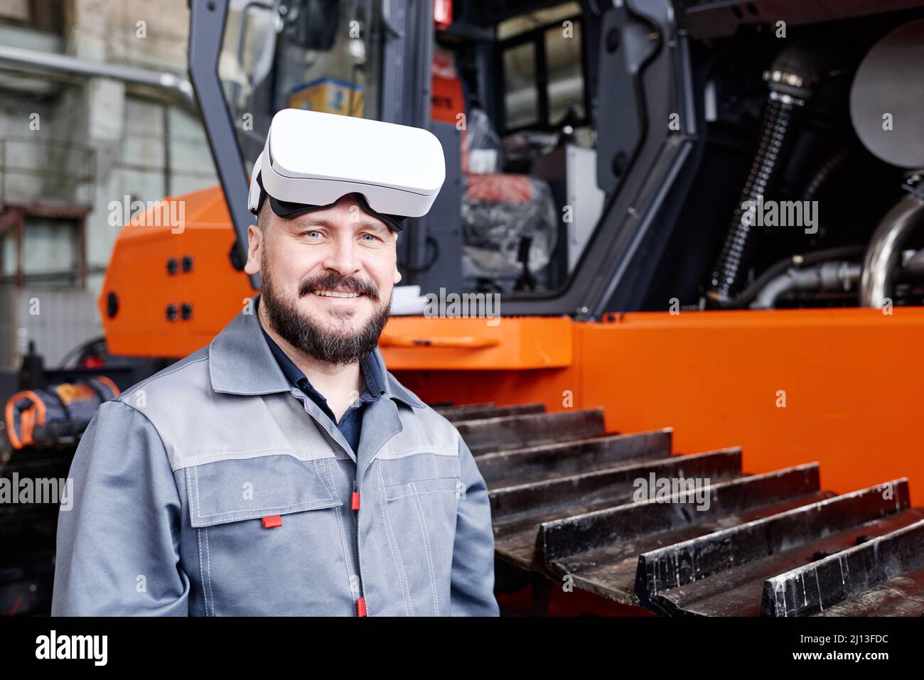 Happy mature worker of factory wearing vr headset and workwear looking at cameras while standing by industrial machine Stock Photo