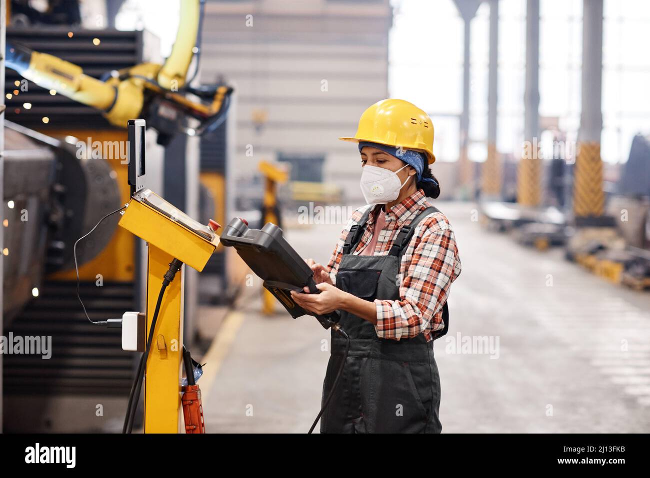 Young female technician in protective workwear using remote switch to control industrial machine while standing in large workshop Stock Photo
