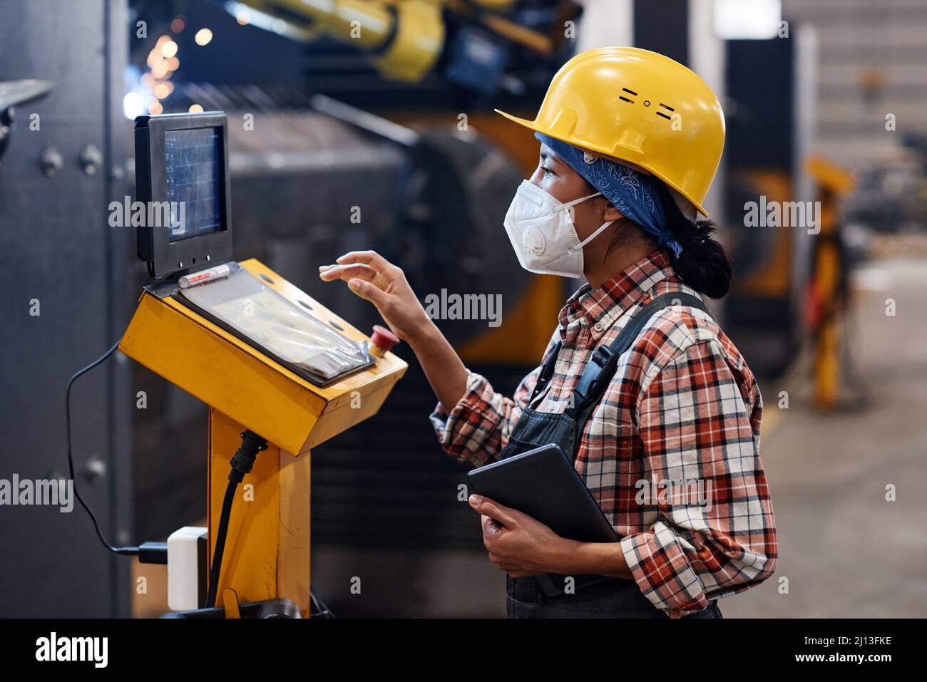Young woman in protective helmet, respirator and coveralls pointing at document while looking at data on screen during work Stock Photo