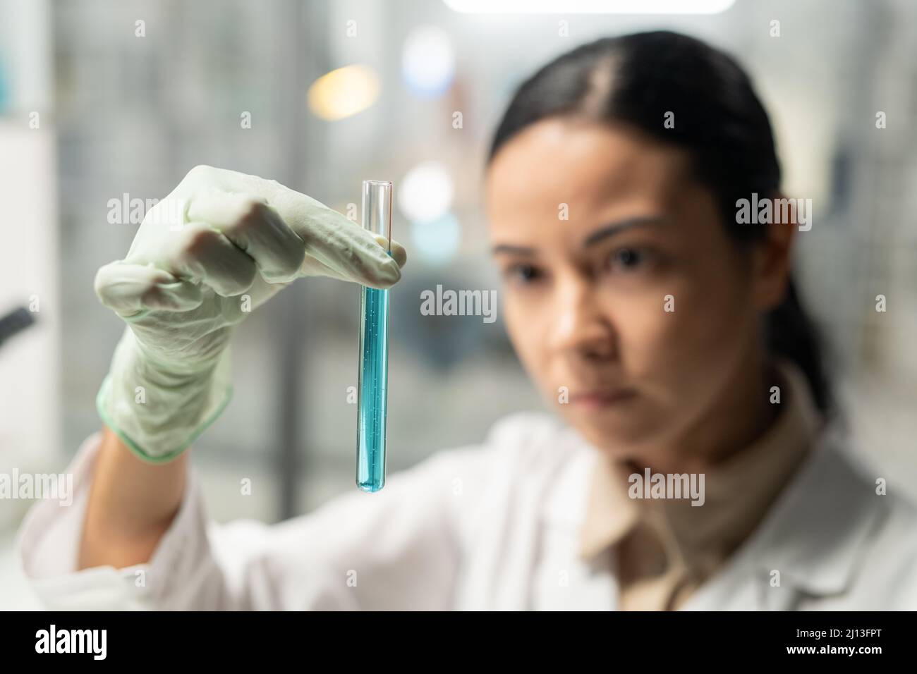 Gloved hand of young female scientist in lab coat holding flask with blue liquid while carrying out scientific experiment in laboratory Stock Photo