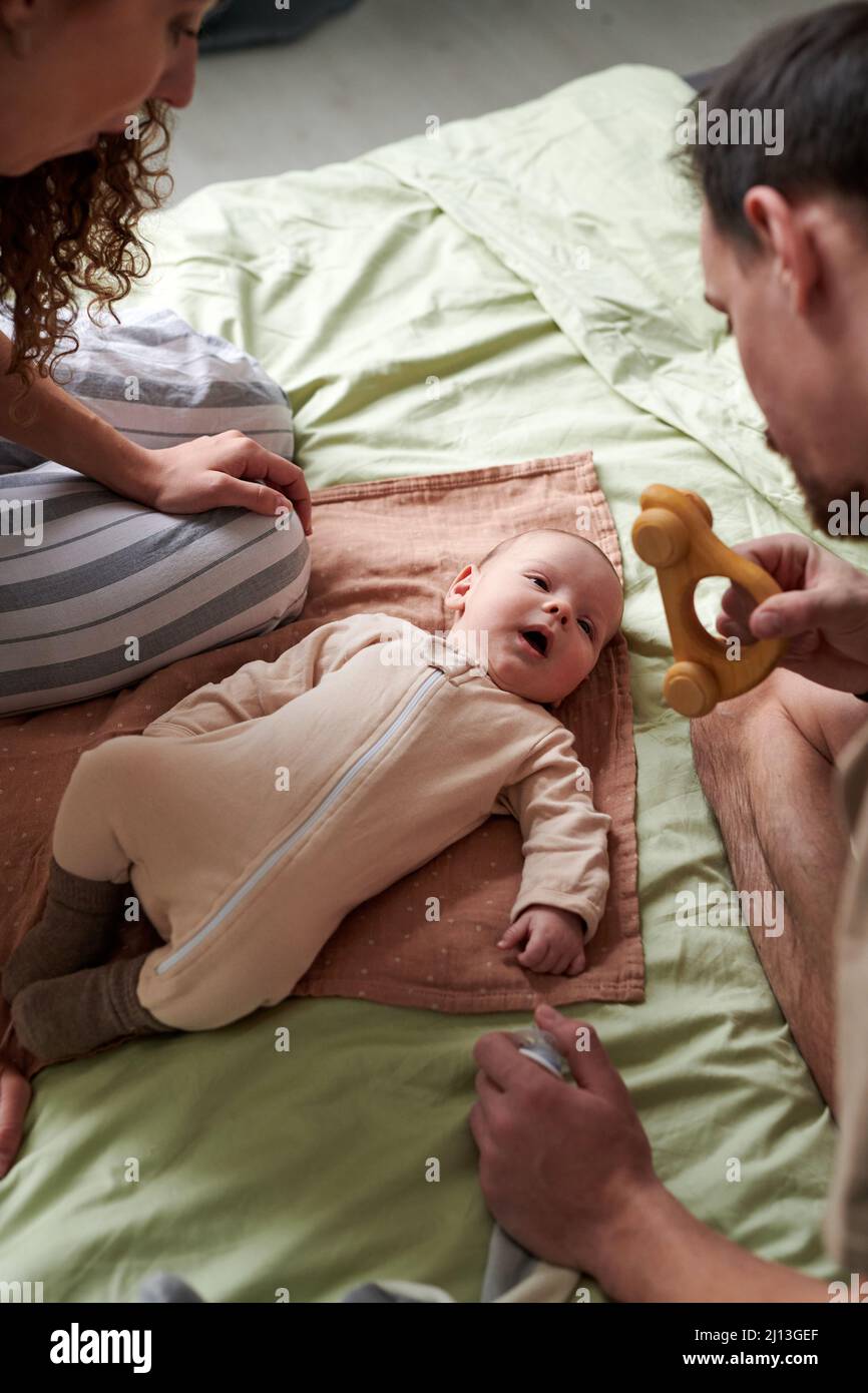 Adorable baby boy lying on double bed and looking at toy held by his father while two young parents bending over him Stock Photo