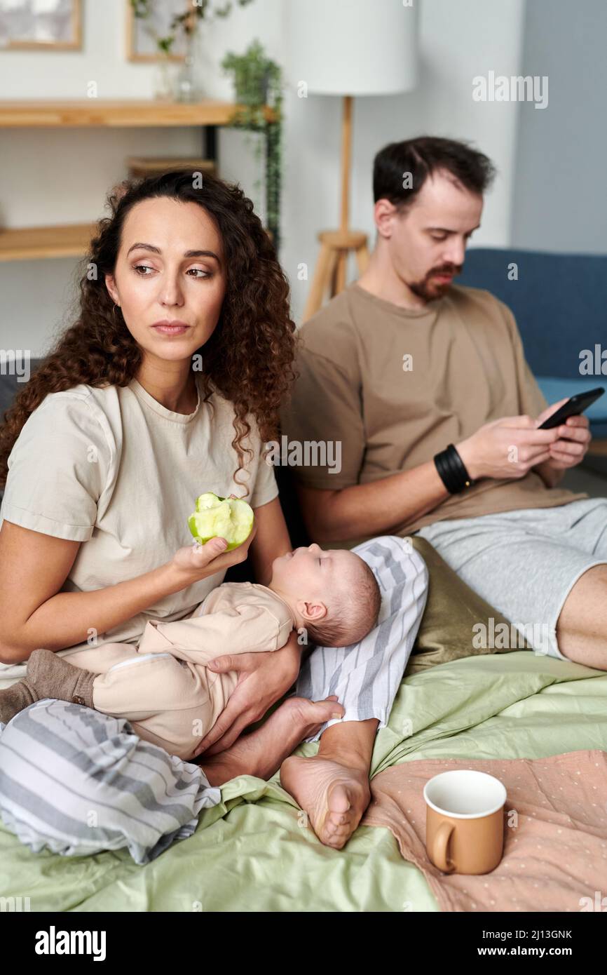 Young serene female with sleeping baby eating fresh green apple for breakfast while sitting on