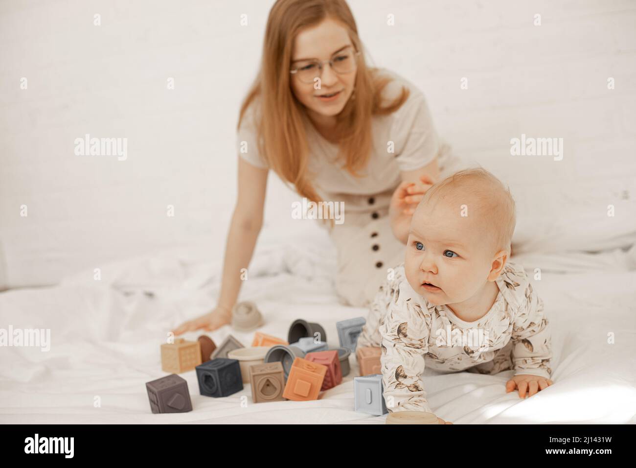 Blond woman teacher in glasses and child playing together with toy cubes on white linen indoor, kindergarten. Baby game  Stock Photo
