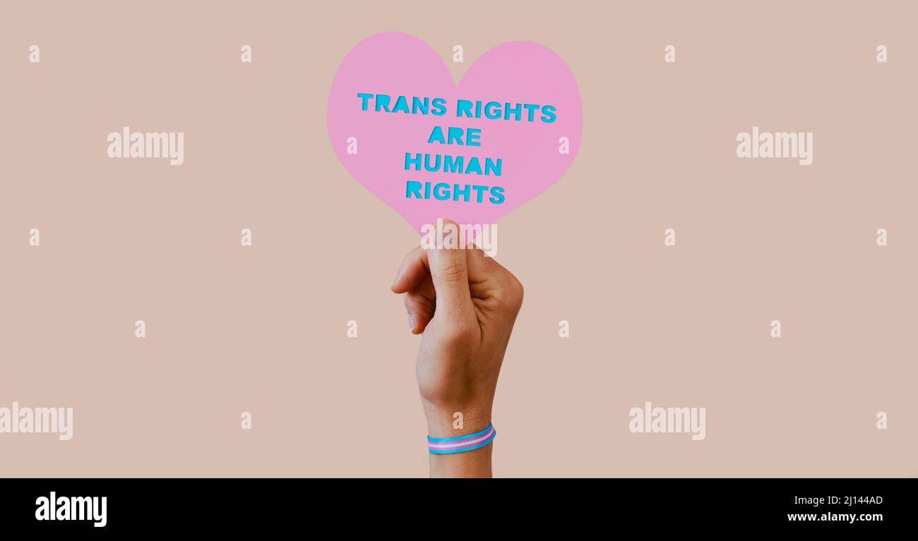 a young caucasian person, wearing a wristband patterned with the transgender pride flag, holds a pink heart with the text transgender rights are human Stock Photo