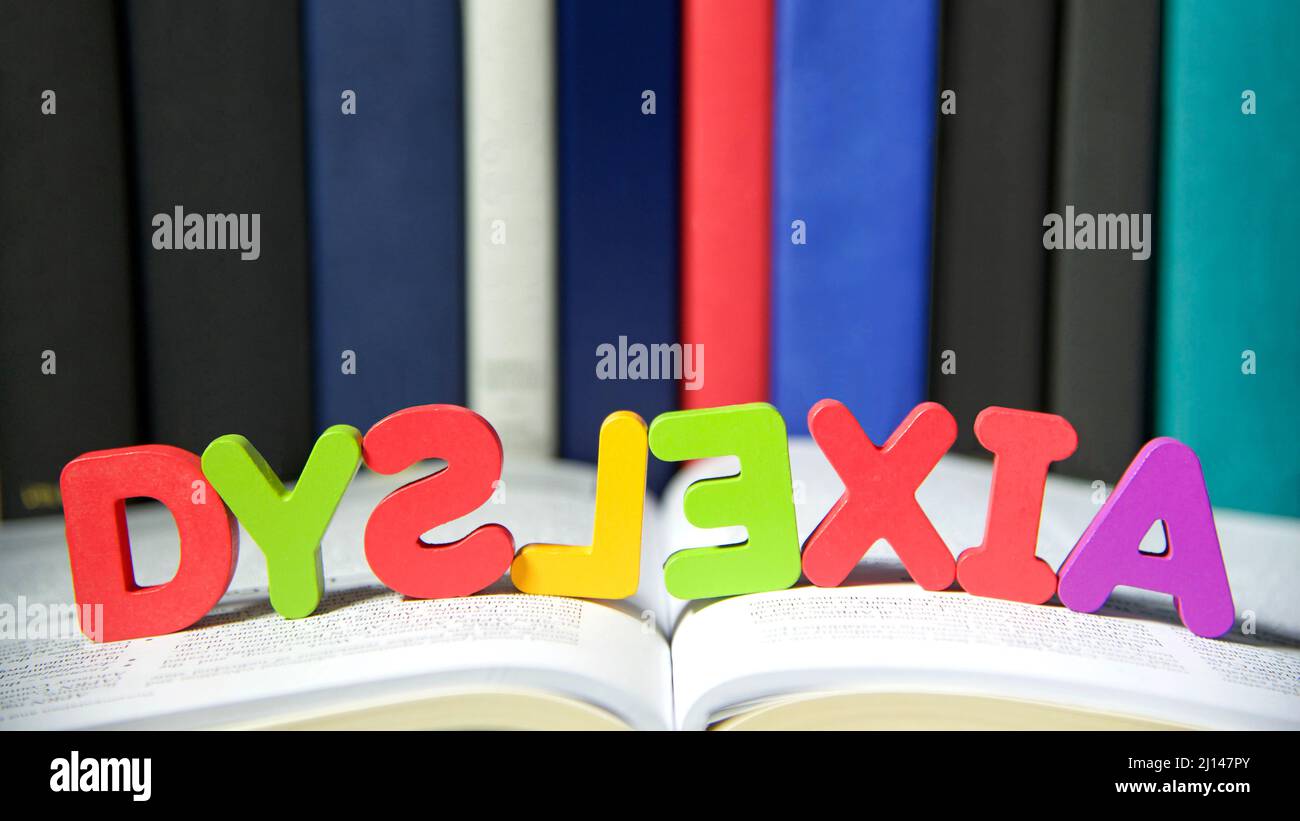 Bright colorful wooden toy blocks on an open book spelling dyslexia. Learning challenges disabilities. S, L and E are backwards. Hard bound books line Stock Photo
