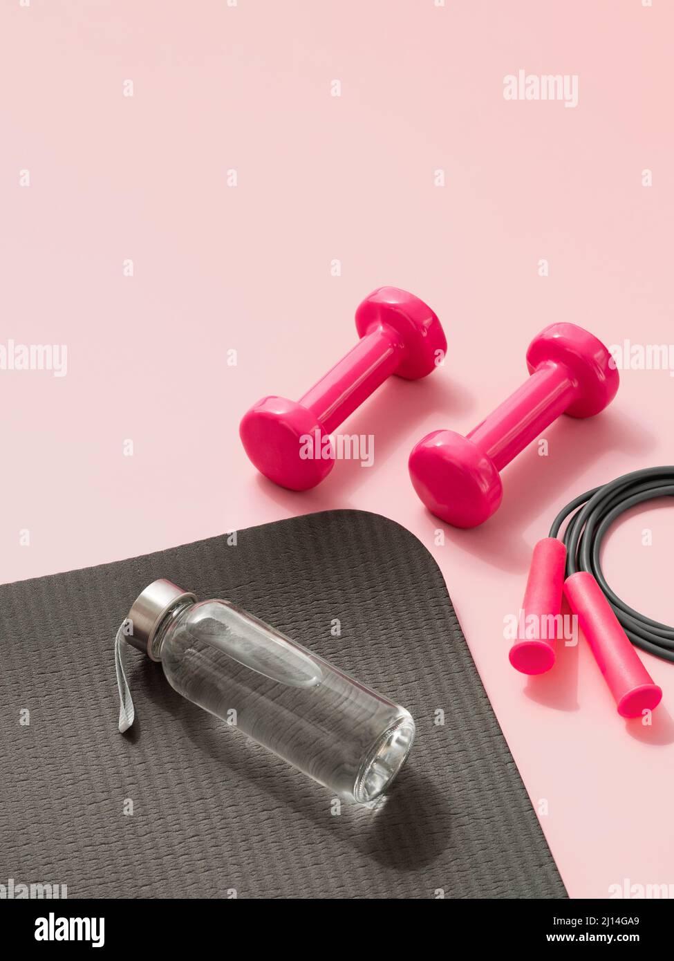 Stylish gray and pink fitness training and gym concept. Perspective view of gray sport mat, water bottle, pink dumbbells and skipping rope on pink background. Set for pilates, fitness with copy space Stock Photo