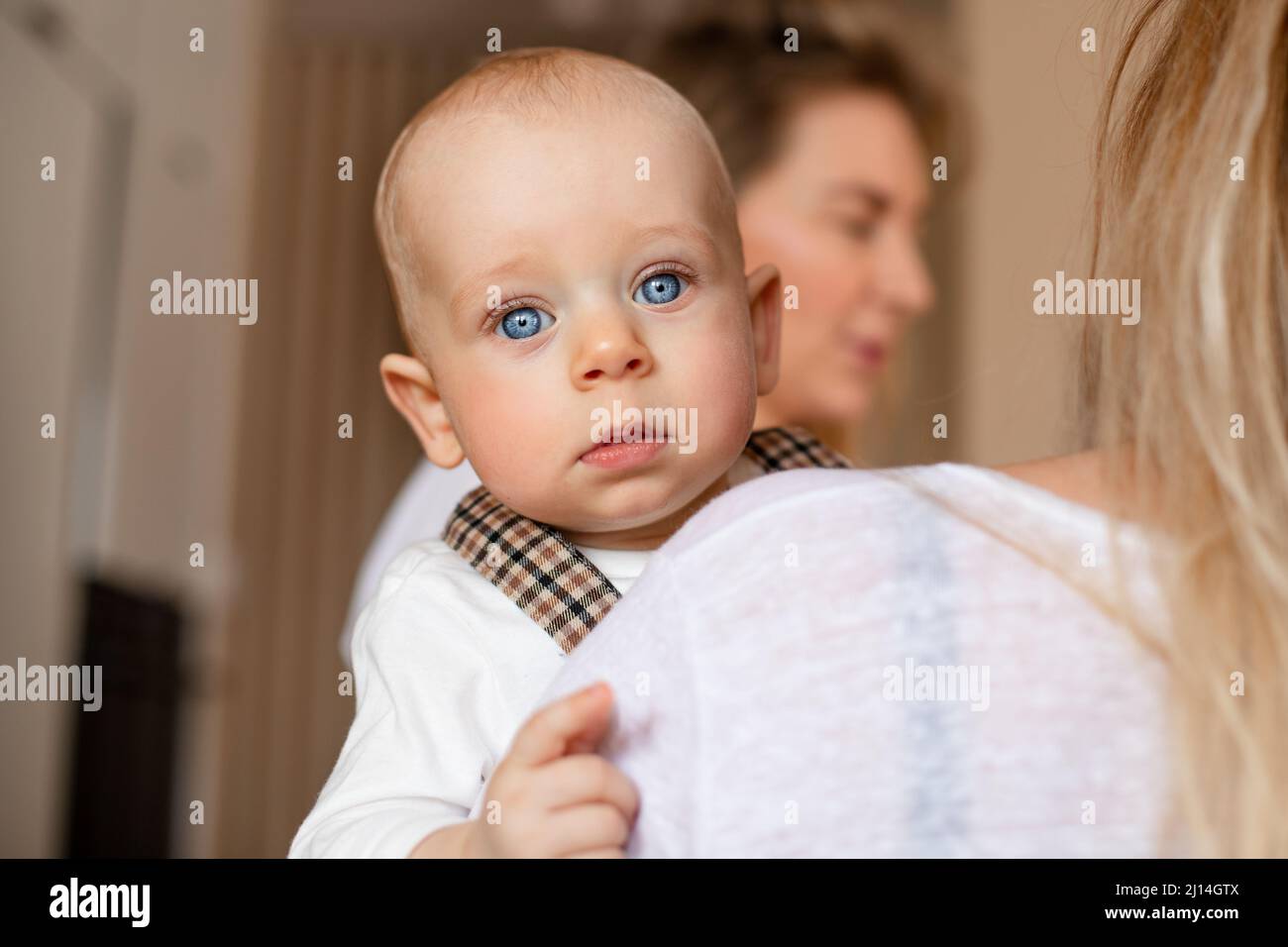 Cute blonde baby with big blue eyes looking at camera. Mom holds her toddler in her hands Stock Photo