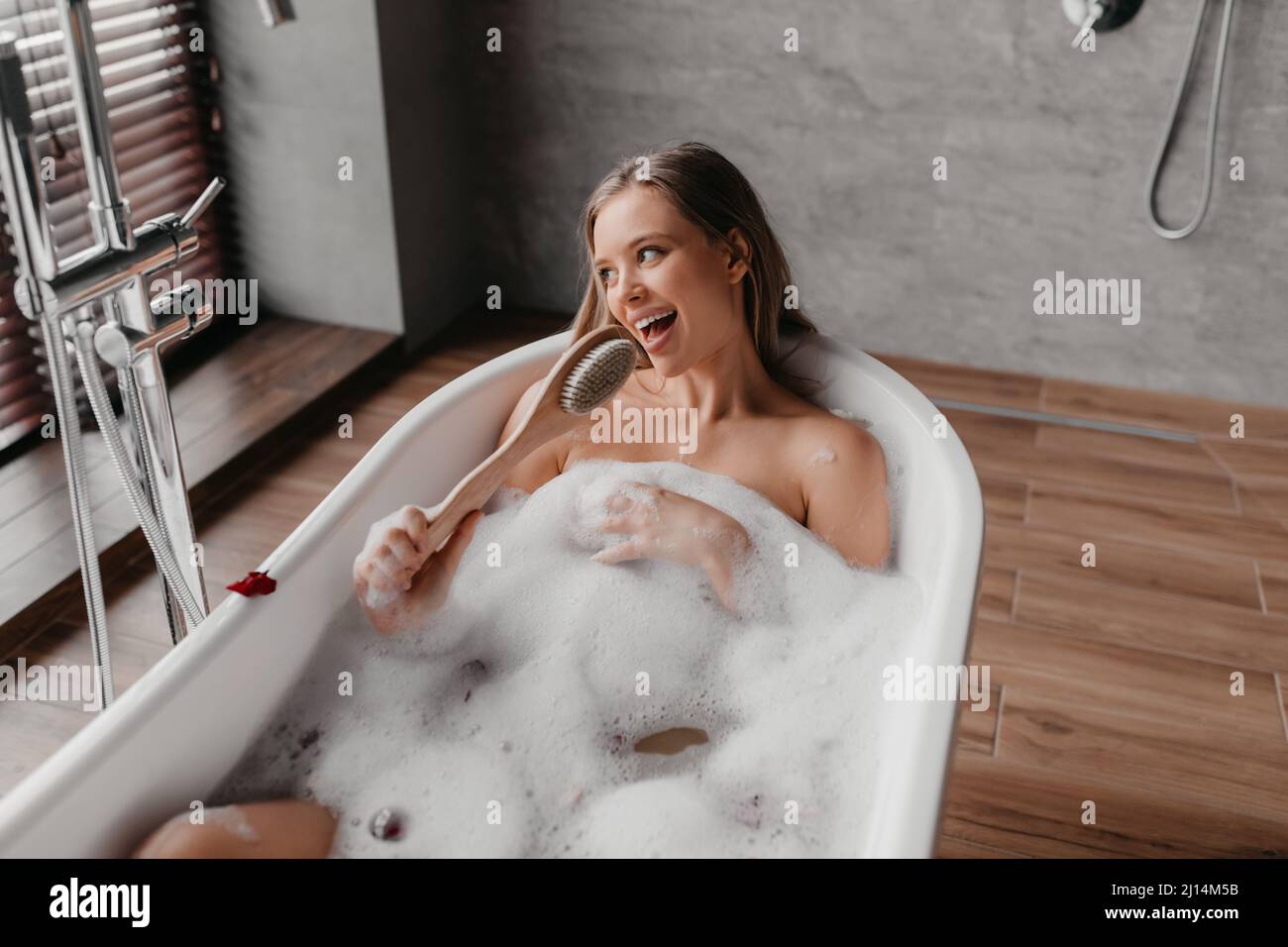 Happy woman singing song holding body brush as microphone, resting in foamy bath at home Stock Photo