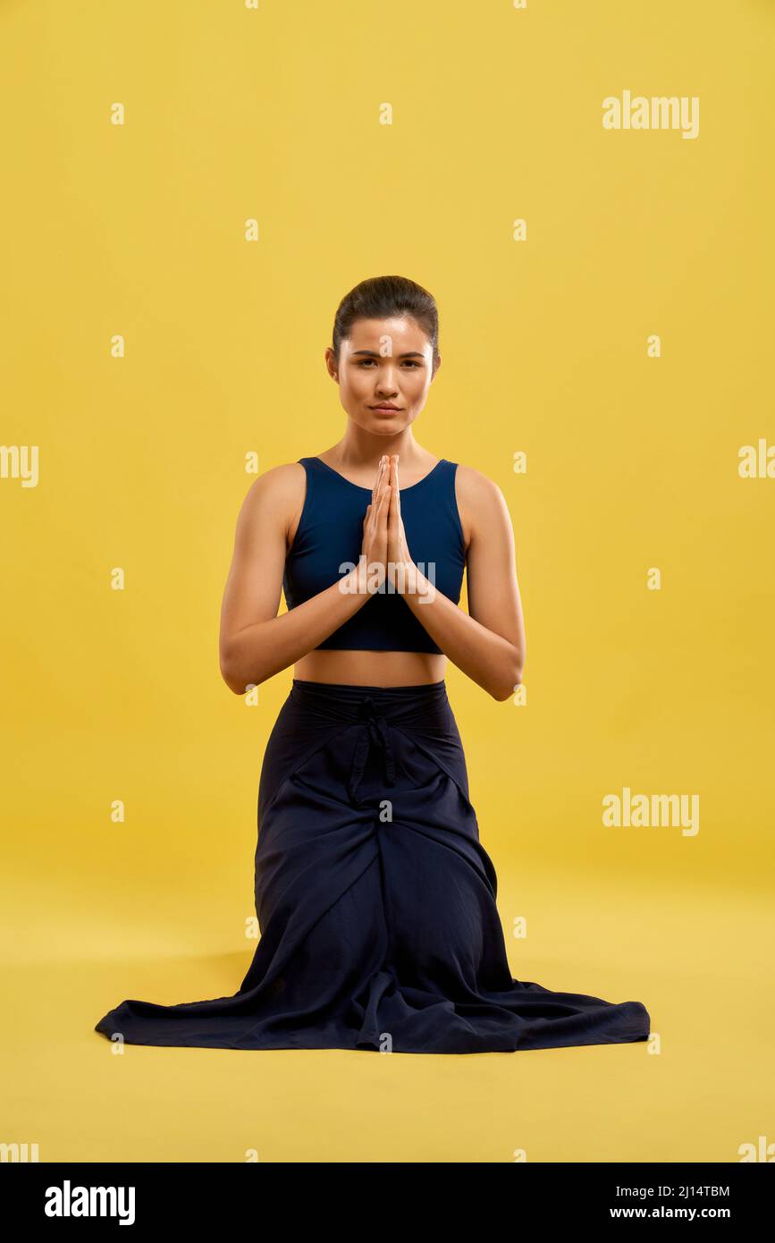 Front view of woman standing on knees, holding prayer hands, looking at camera. Slim girl wearing sport suit, practicing yoga pose, meditating. Concep Stock Photo