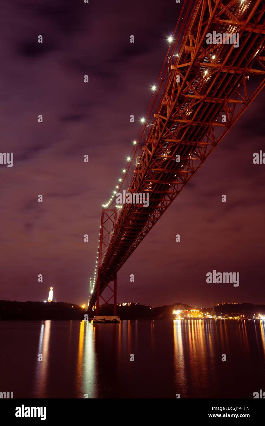 The 25 de Abril Bridge (Ponte 25 de Abril) crossing over the Tagus River (Rio Tejo) at dusk, and the Cristo de Rei - Lisbon, Portugal Stock Photo