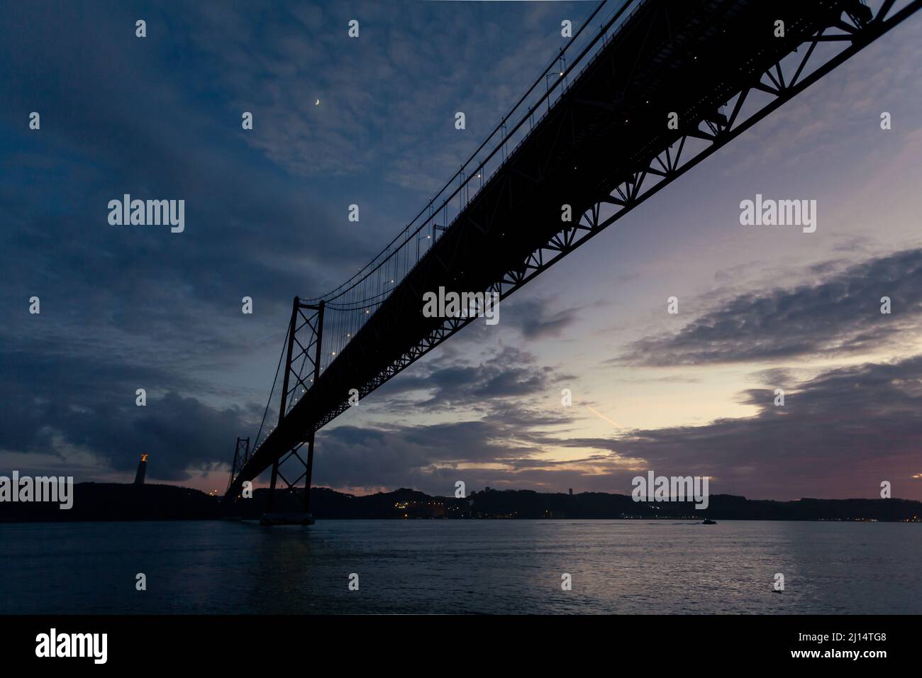 The 25 de Abril Bridge (Ponte 25 de Abril) crossing over the Tagus River (Rio Tejo) at dusk, and the Cristo de Rei - Lisbon, Portugal Stock Photo