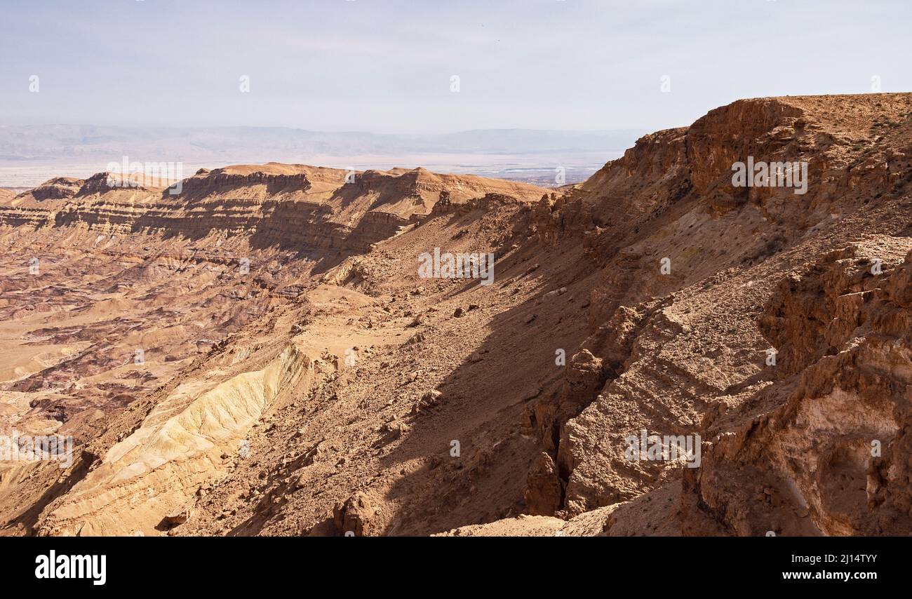 section of the south rim of the Makhtesh Katan Small Crater showing cliffs and colorful sandstone with the Jordan Rift Valley in the background Stock Photo