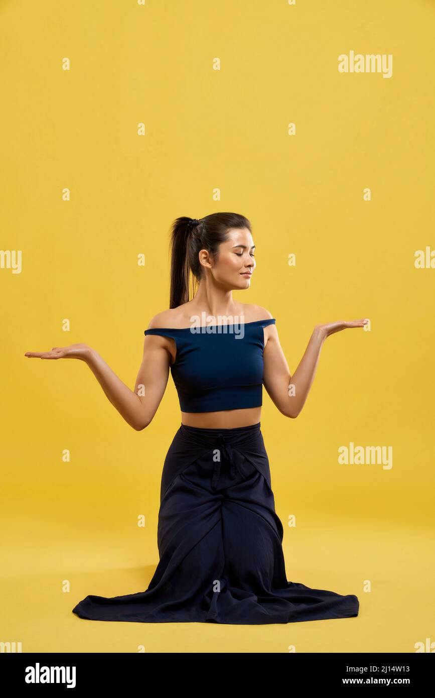 Front view of slim girl with pony tail standing on knees. Pretty woman raising hands, palms, looking down, smiling, yoga pose practicing. Concept of n Stock Photo