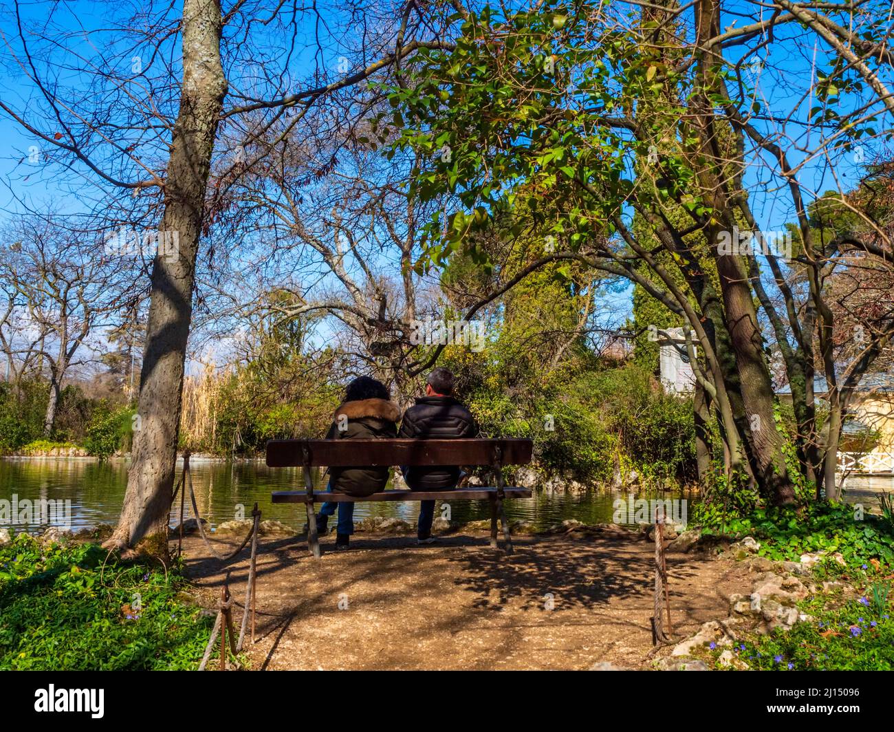 A heterosexual couple sitting under the trees on a public bench in a park in Madrid. Stock Photo