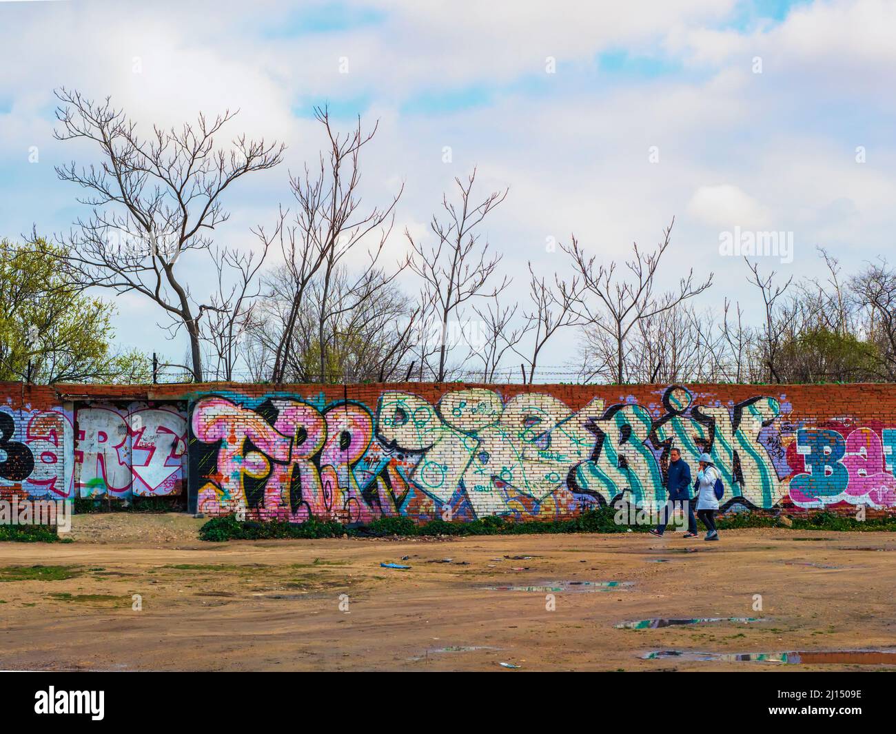 A heterosexual couple walks through an open field in front of a wall with graffiti. Stock Photo
