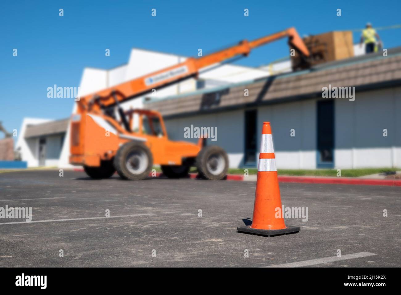 Orange traffic cone on asphalt parking lot near hydraulic forklift where a building roof is being repaired. Stock Photo