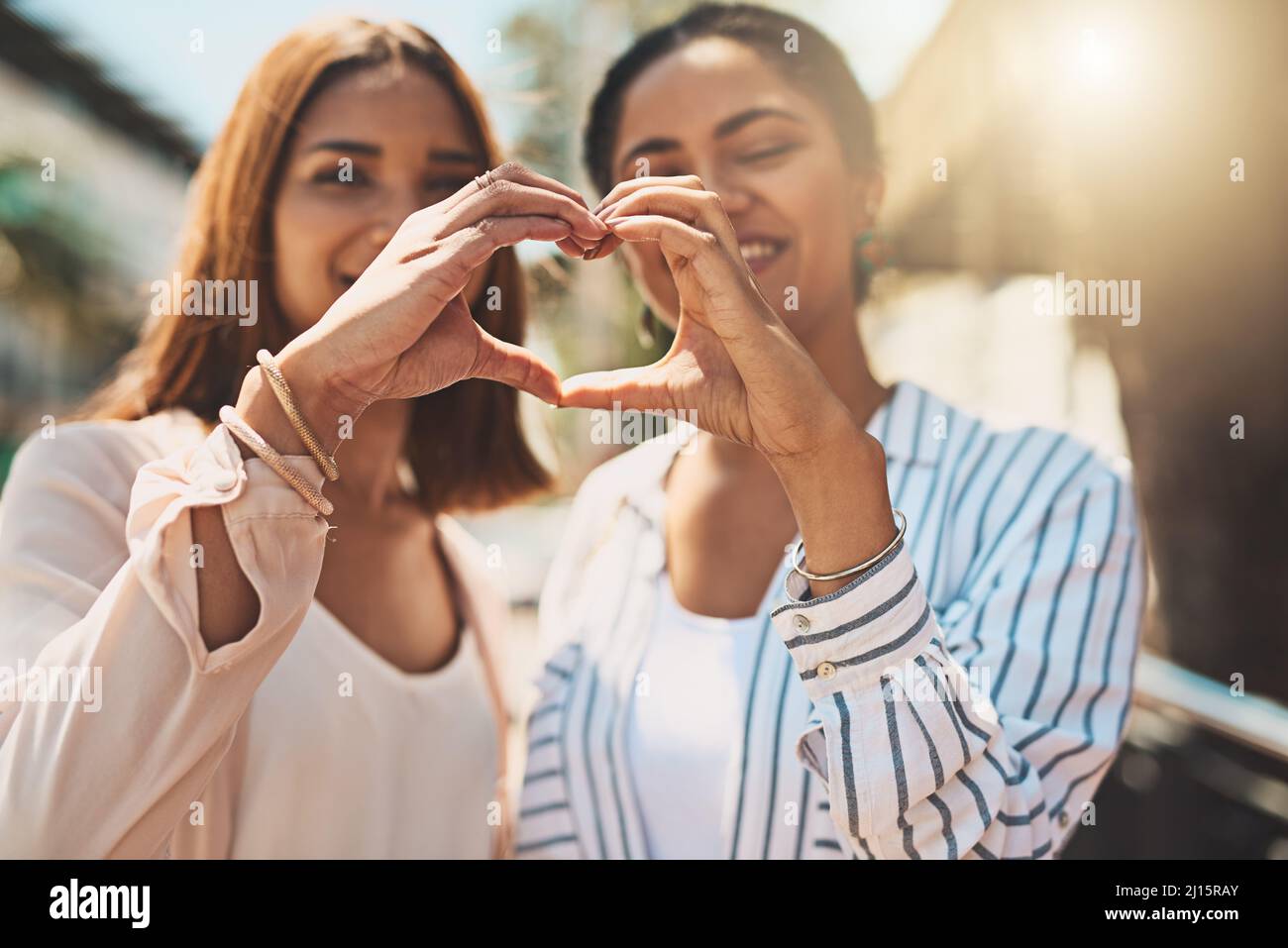 Love keeps your friendships strong. Shot of two cheerful young women forming a heart shape with their hands together while standing outside during the Stock Photo