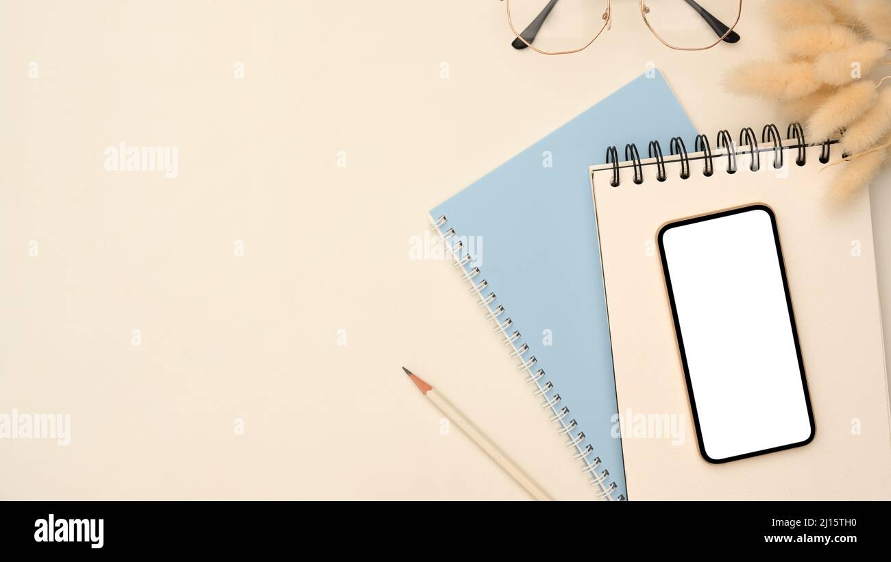Overhead shot, Modern workspace with smartphone blank empty screen mockup on spiral notepad, eyeglasses, pencil and copy space on white background. Stock Photo