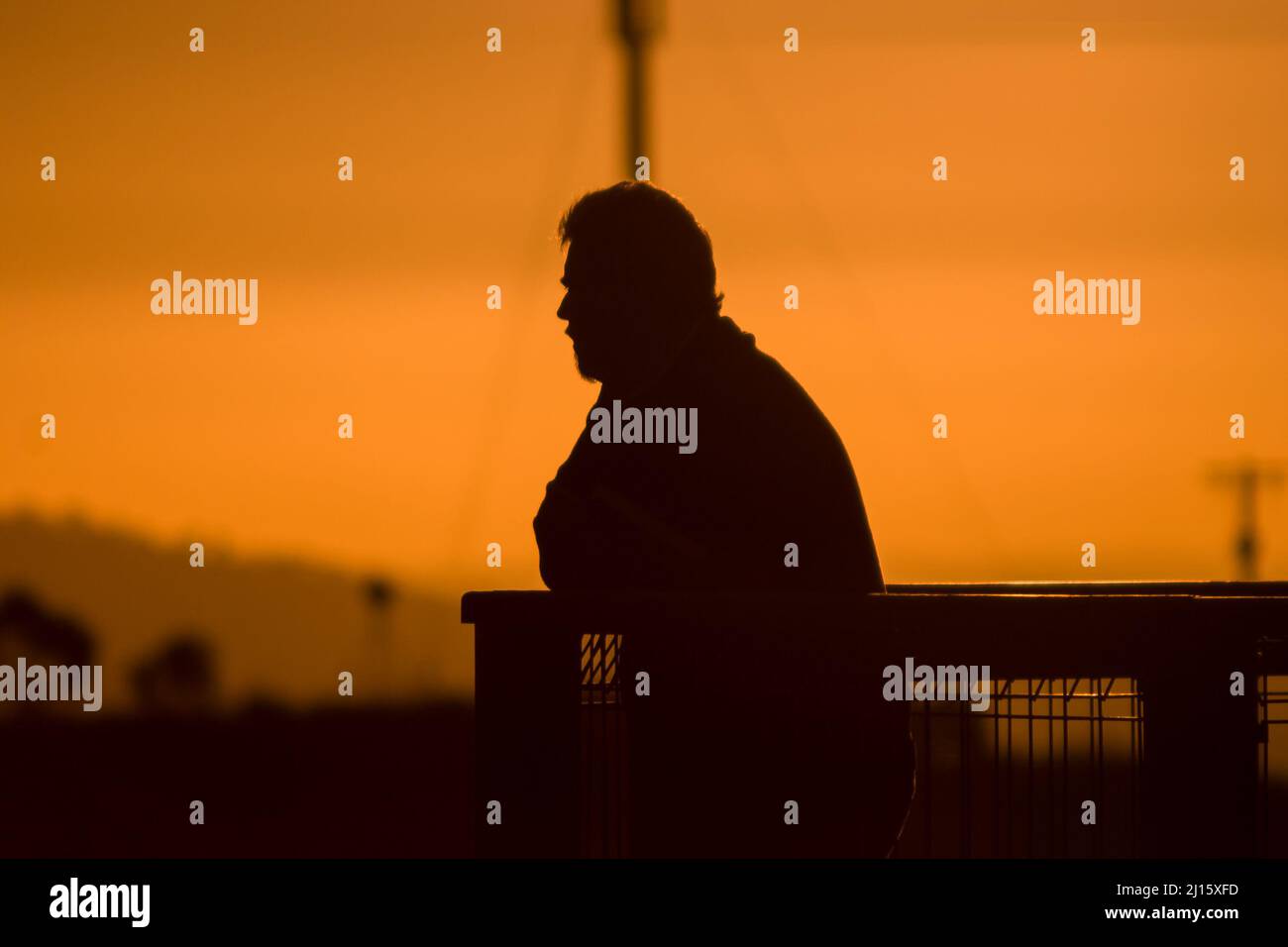 Silhouette of man with beard at sunset leaning on fence looking out at view Stock Photo