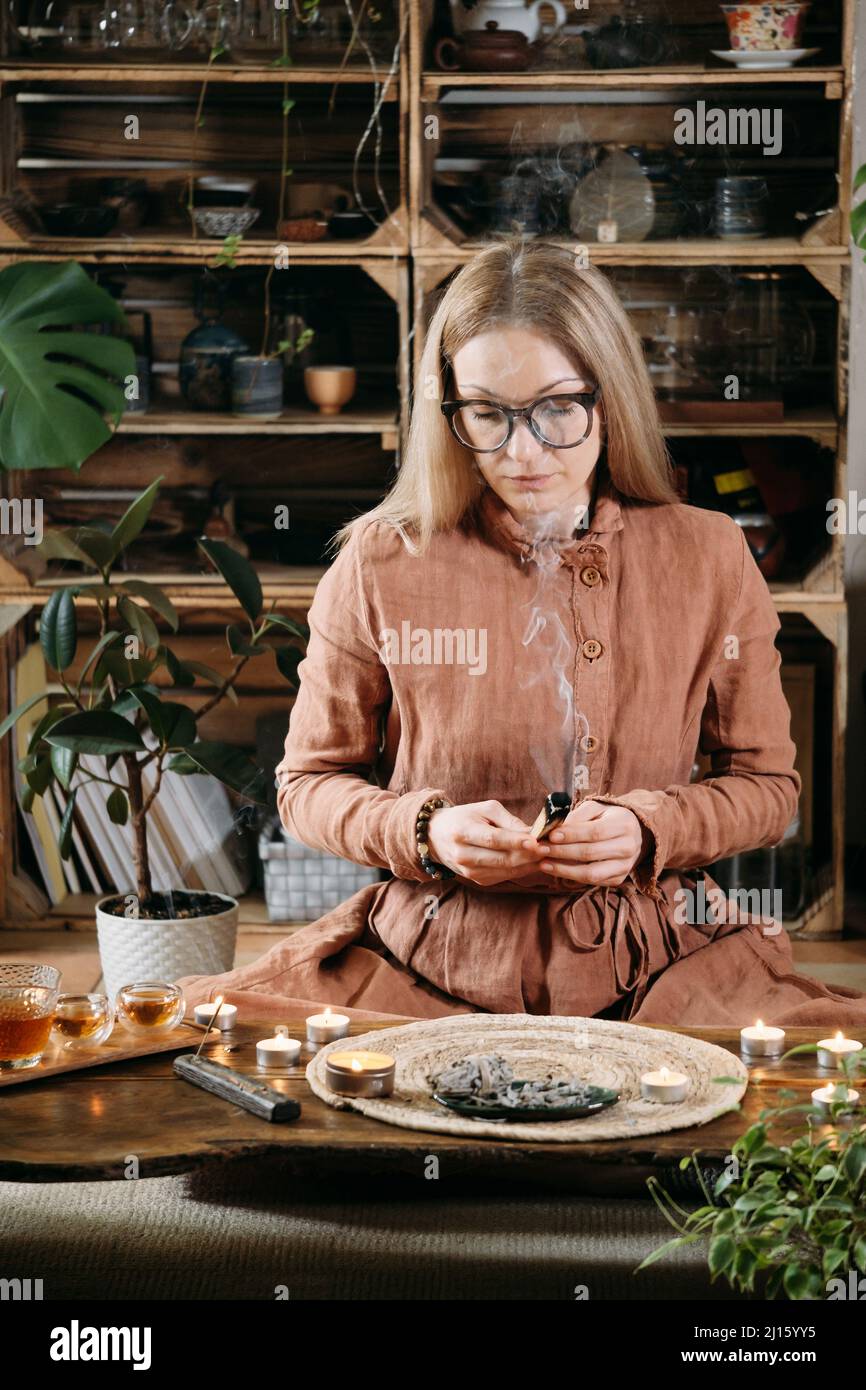 Woman meditating with palo santo in studio, cleaning space with smoke. Adult person using burning spiritual incenses at home Stock Photo