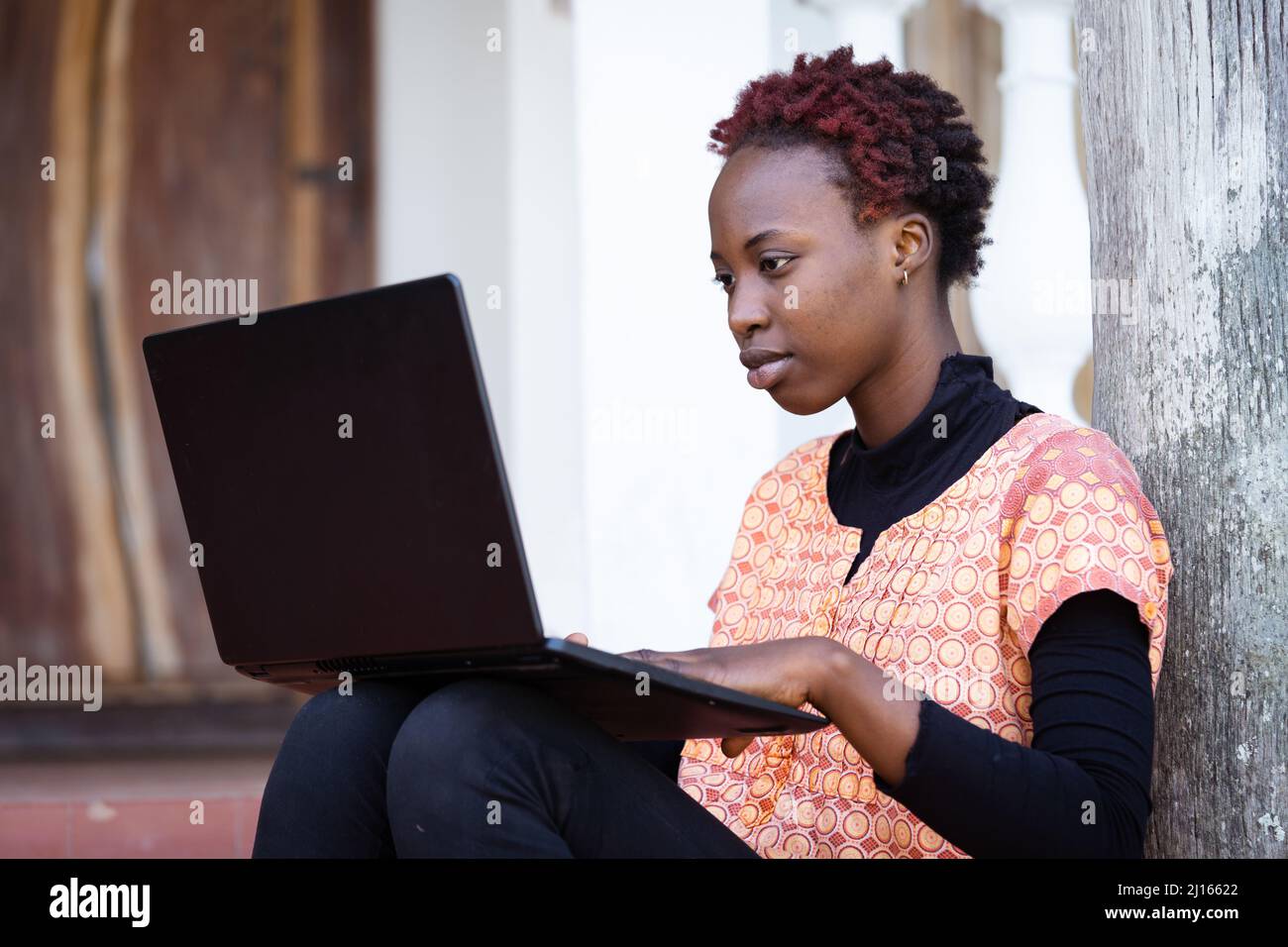 Concentrated African teenager sitting on the porch with her laptop on her legs preparing online for her high school exam; education during COVID pande Stock Photo