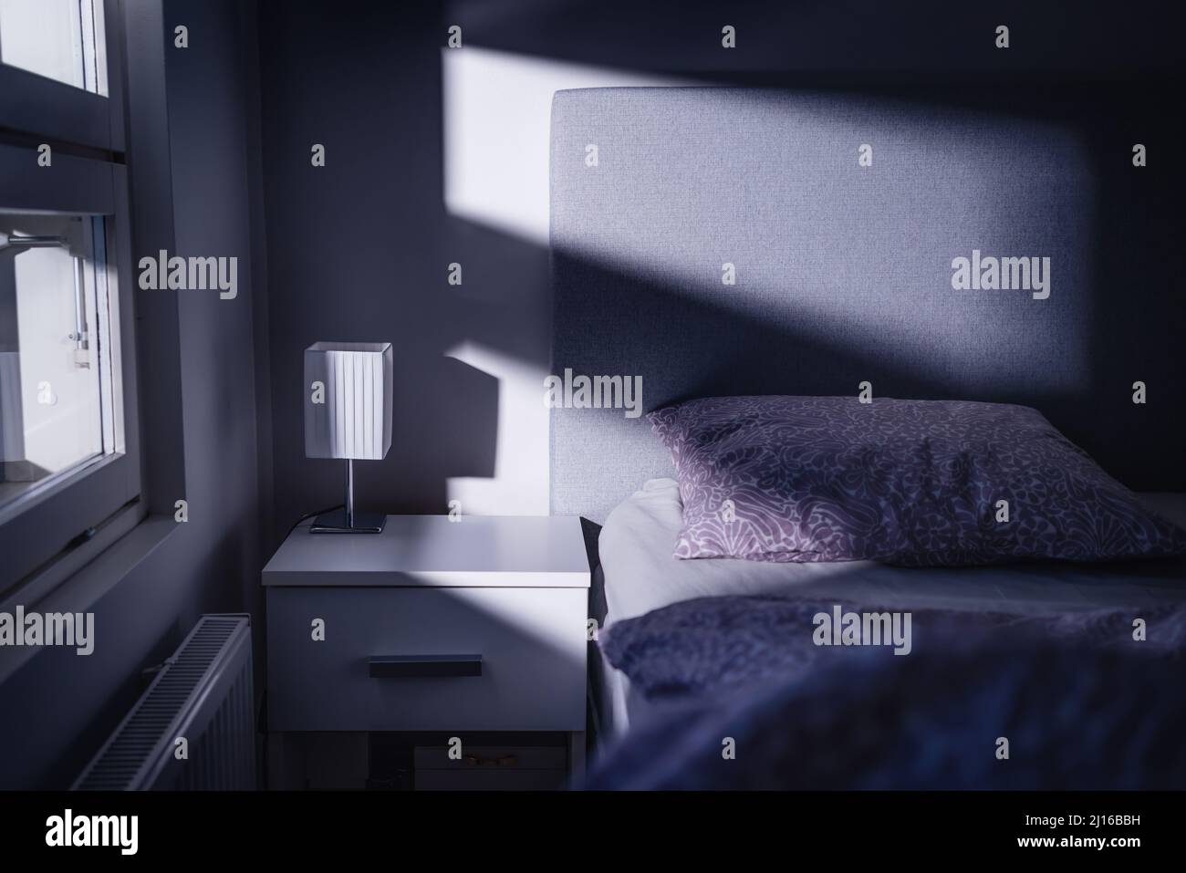 Bed at night in dark bedroom. Blue light and moonlight from window. Pillow, sheet and duvet ready for sleeping. Bedside table and nightstand. Stock Photo