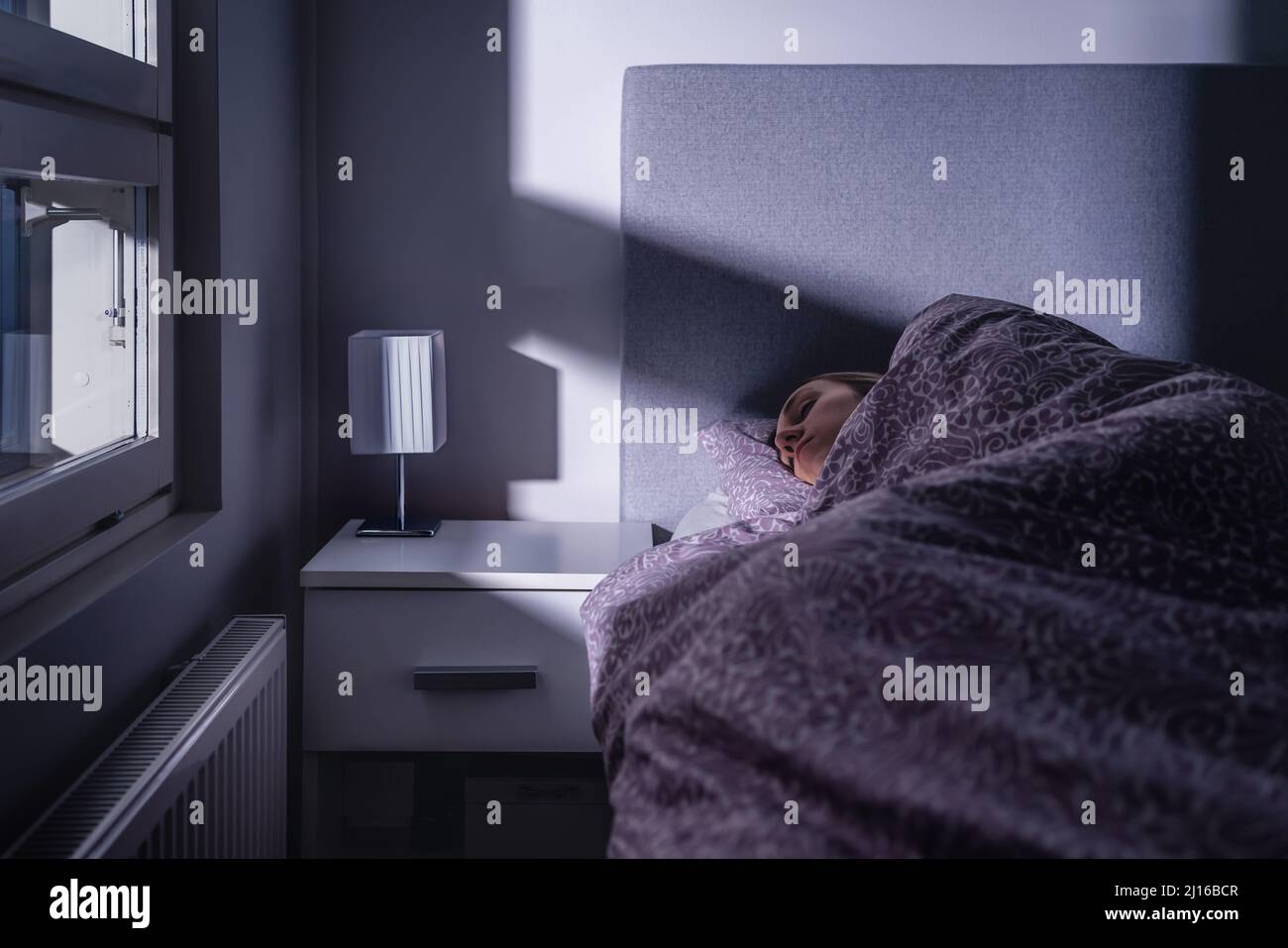 Sleeping woman in bed at night in dark bedroom. Person resting. Asleep under cover and blanket. Cold room at home. Tired lady in peaceful dream. Stock Photo