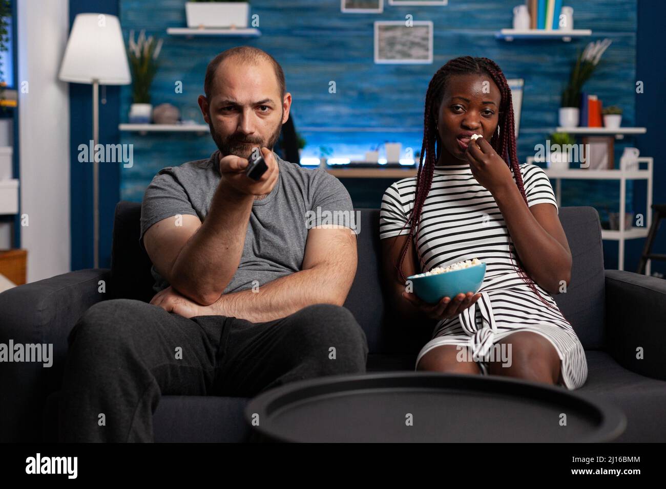 Caucasian man and african american woman watching TV while sharing a bowl of popcorn sitting on sofa at home. Stressed husband angrily switching television programs looking for a movie. Stock Photo