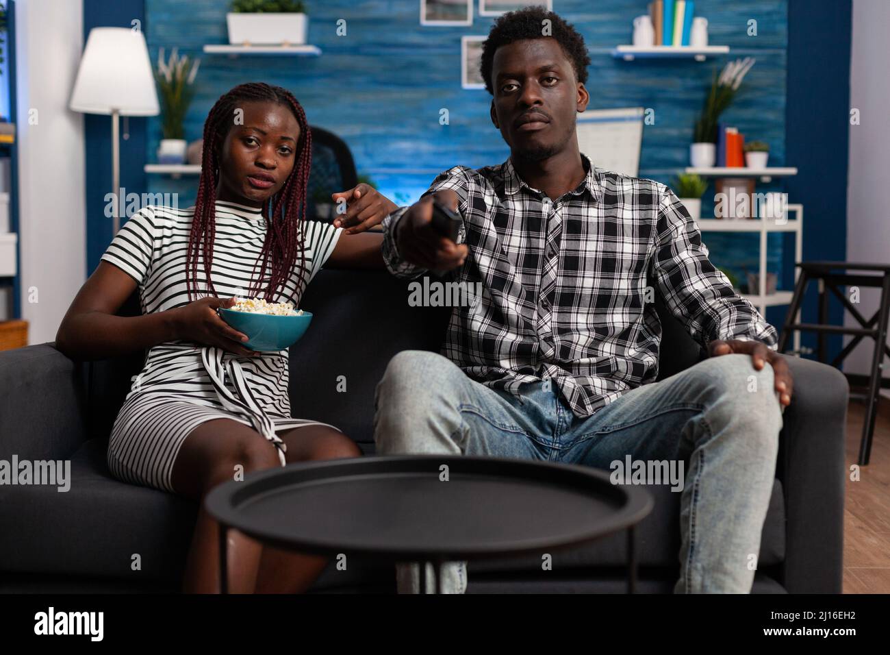African american woman eating popcorn on sofa while young man changing channels on TV using remote. Adult people enjoying being together while watching movies and eating snacks at home. Stock Photo