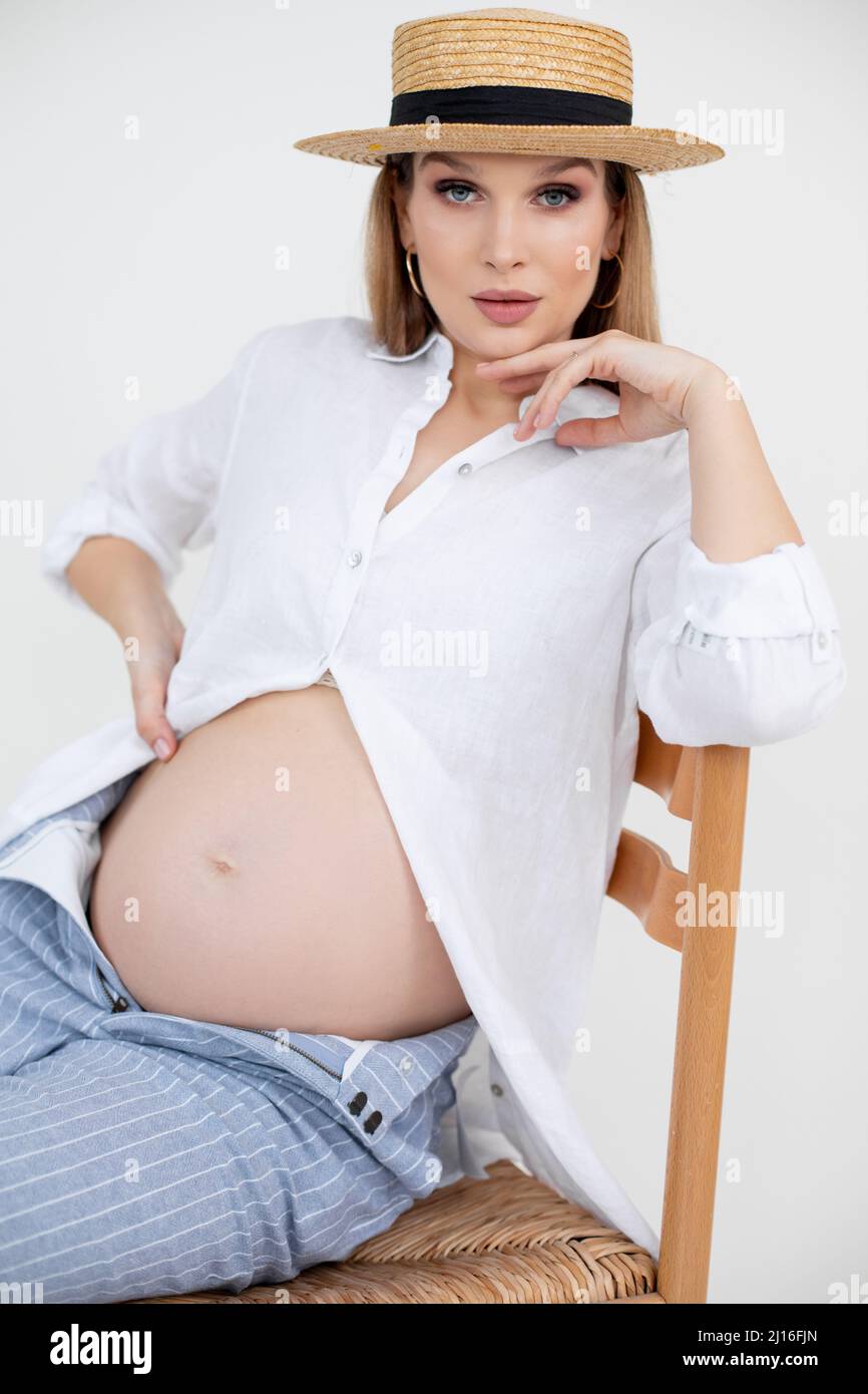 Portrait of young pregnant woman with long dark hair and makeup wearing golden earrings, white shirt and straw hat. Stock Photo