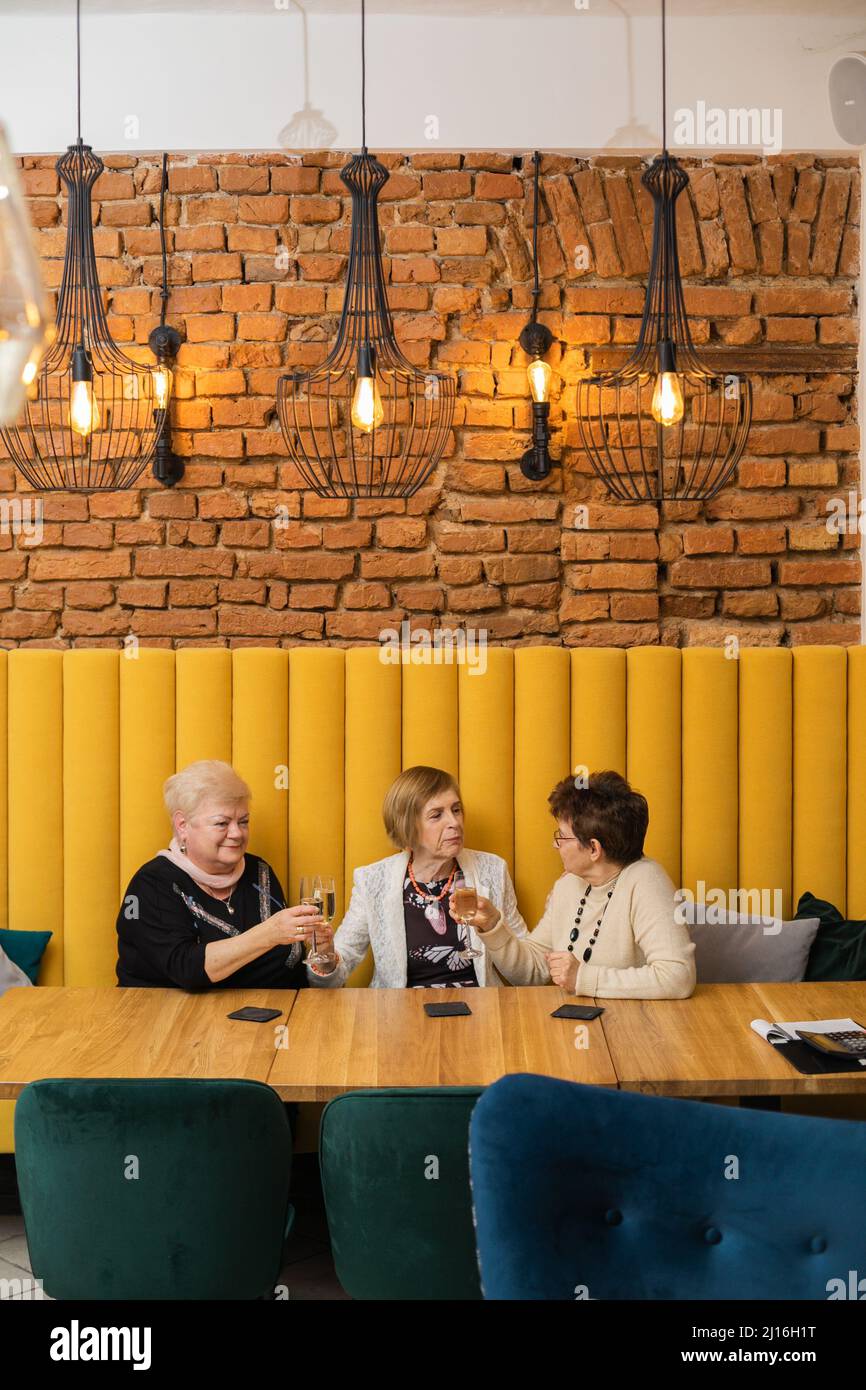 Three smiling elderly women with short dark and fair hair clink glasses of champagne sitting celebrating at restaurant. Stock Photo