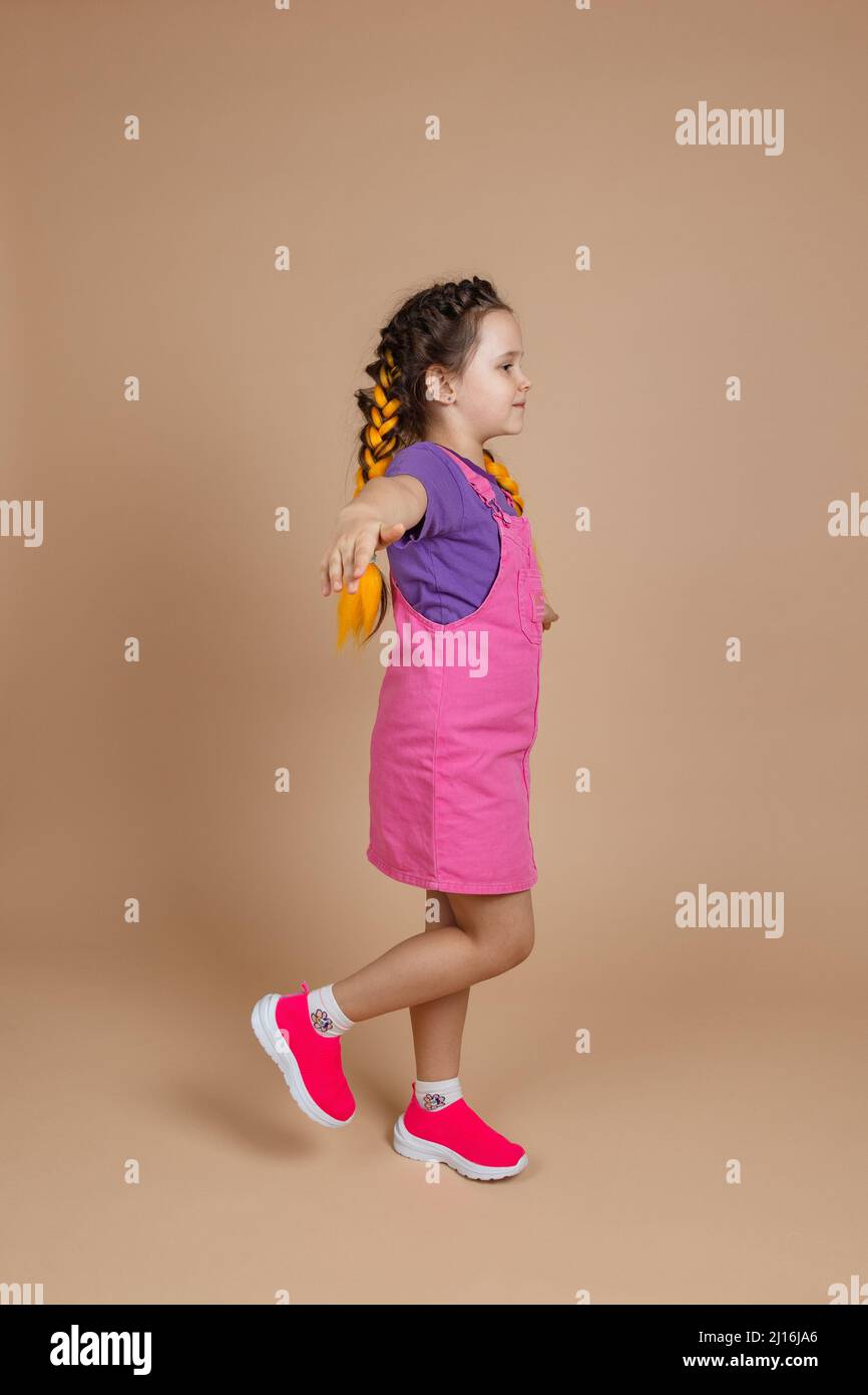 Happy cute little girl having yellow kanekalon braids, balancing on one leg with hands raised and put to sides looking ahead wearing pink jumpsuit and Stock Photo