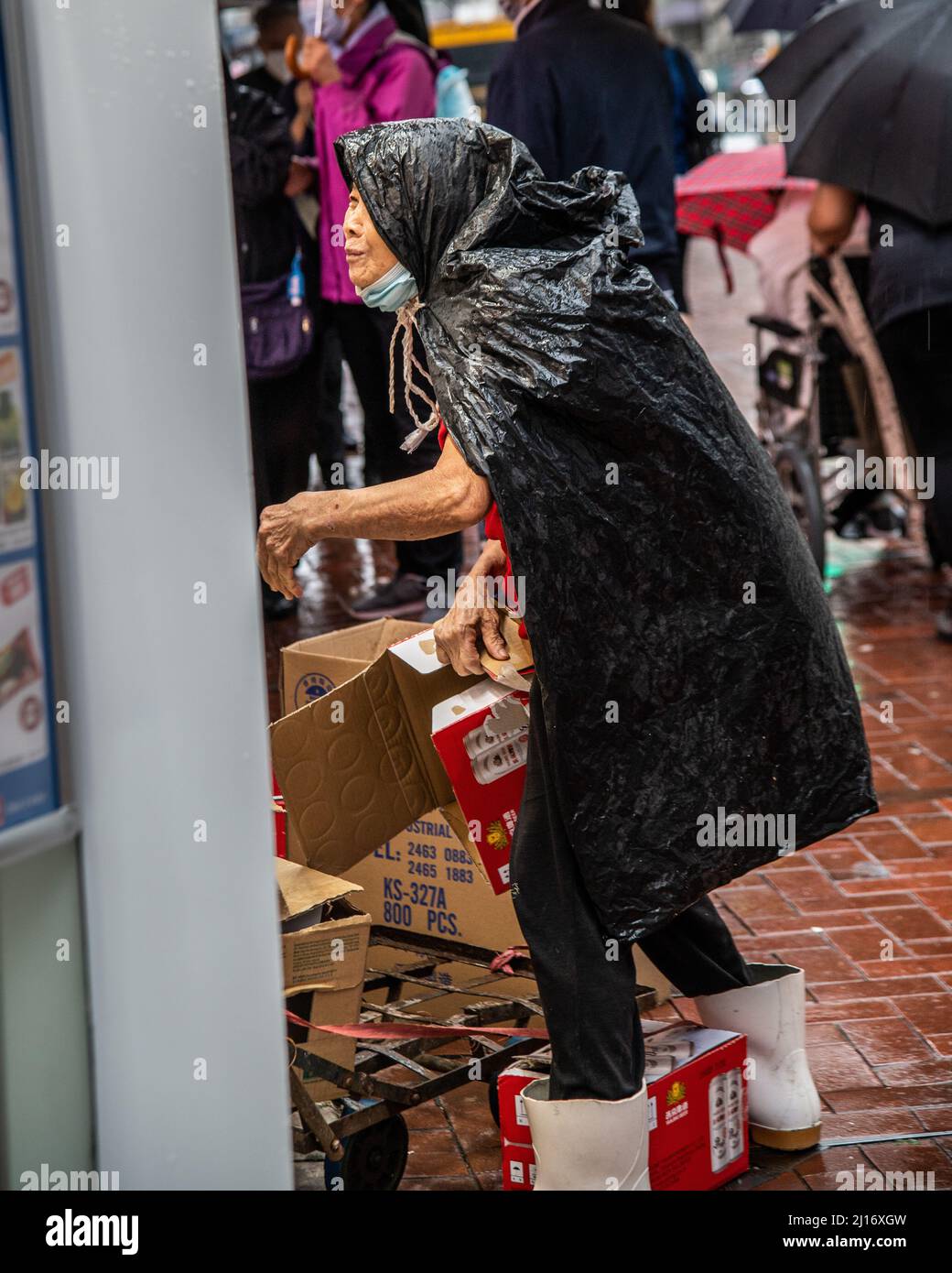 An elderly woman covered with a plastic to protect her from the rain is seen collecting cardboard as a northern monsoon brings cooler weather and rain to Hong Kong. A rainy, spring day in Hong Kong. Stock Photo