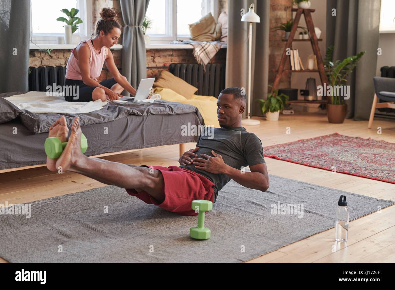 Horizontal shot of sporty young African American man wearing comfortable clothes doing morning exercise at home using dumbbell Stock Photo