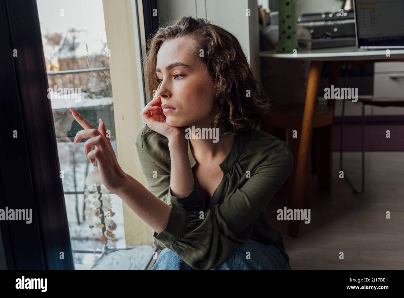Sad woman with head in hand sitting by glass window at home Stock Photo