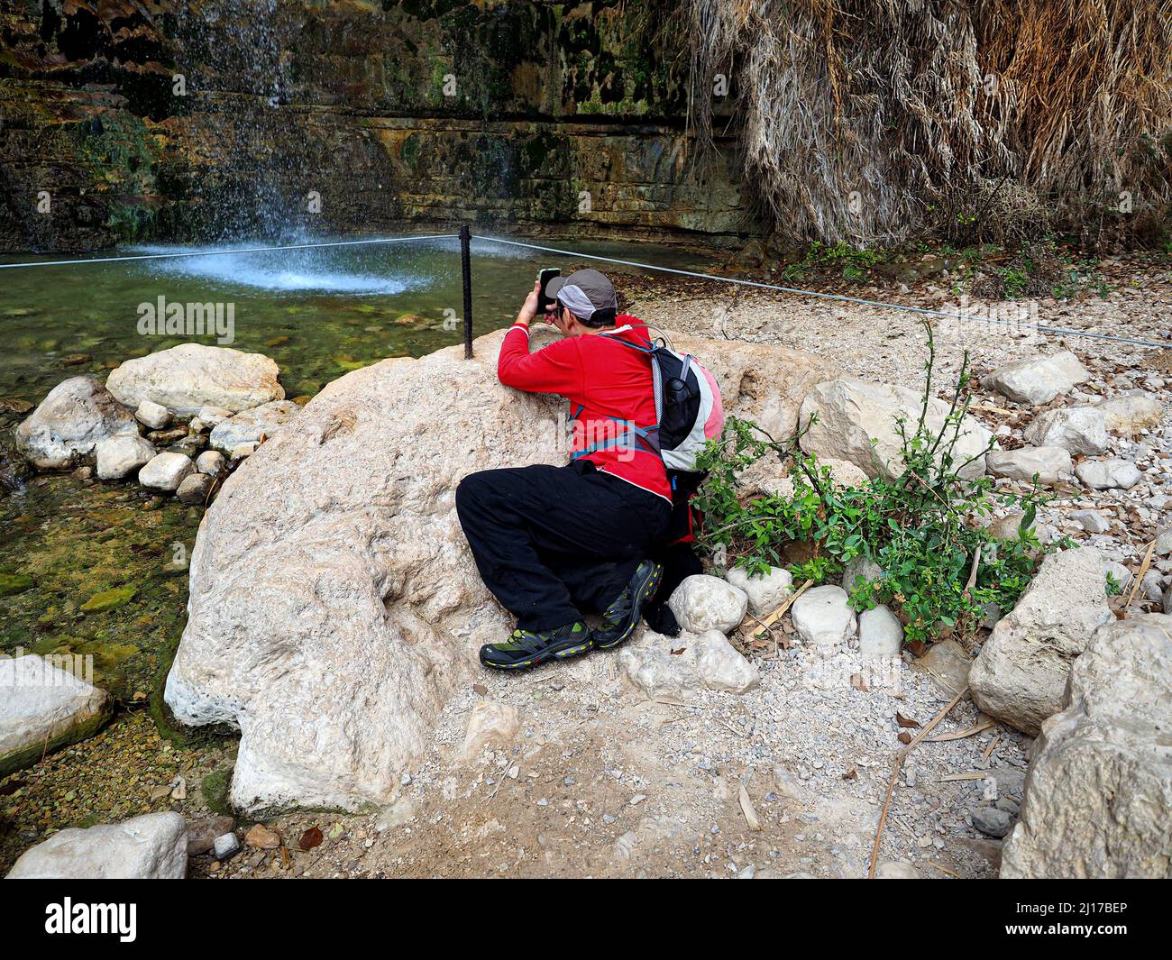 A man sits by a waterfall and takes photos on a smartphone. David Falls in Ein Gedi Nature Reserve. Stock Photo