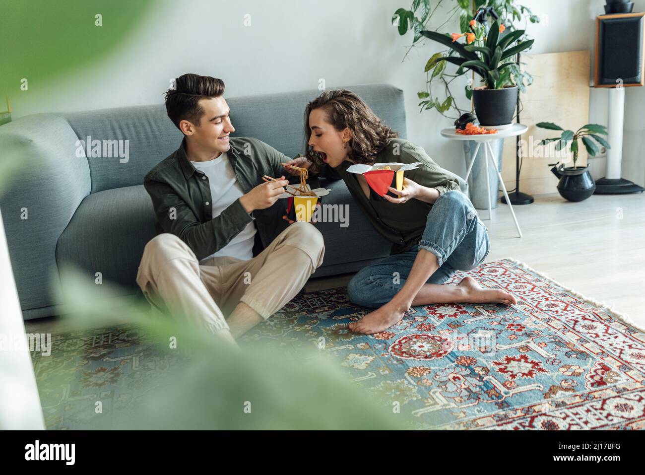 Happy young man looking at girlfriend eating noodles in living room Stock Photo